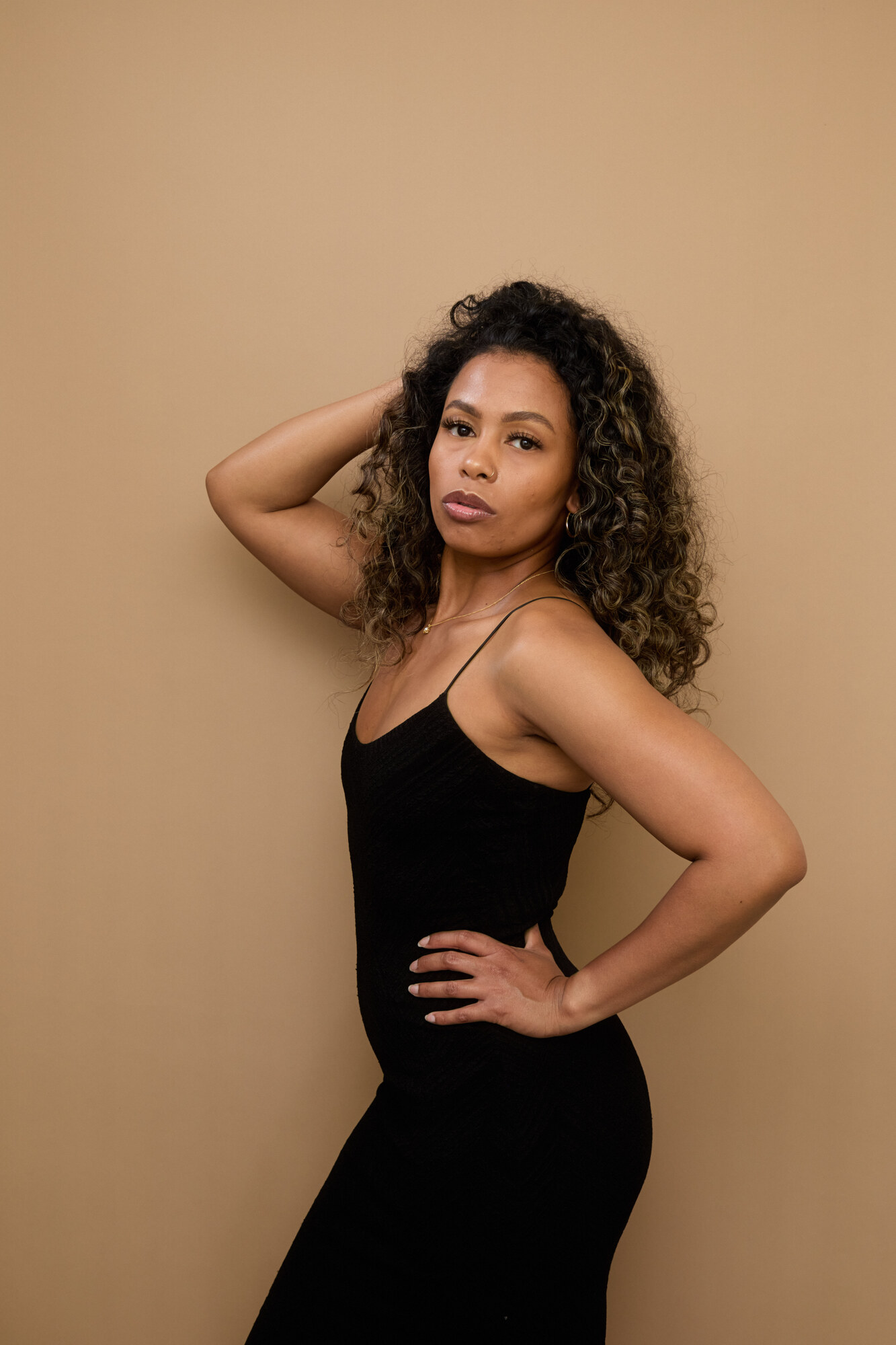 Tash Williamson in a side-angle studio portrait against a plain warm tan backdrop, wearing a black slip dress with thin straps, one hand raised behind her head tousling her loose curly hair, looking back over her shoulder at the camera.