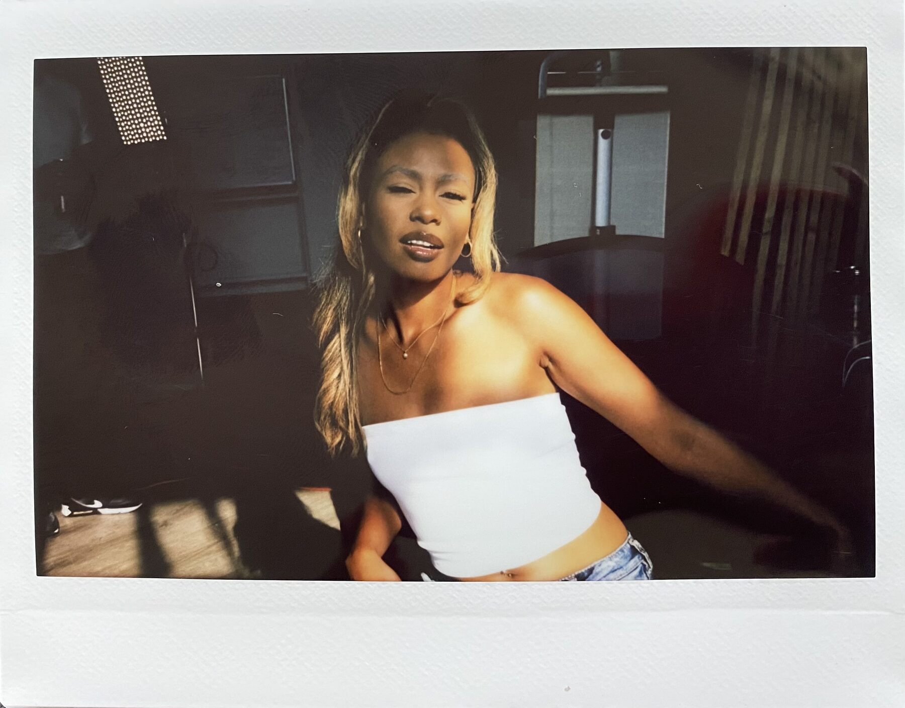 Tash Williamson in a warmly lit Instax-style photograph, leaning toward the camera with lips parted and one arm reaching out of frame in a soft motion blur, wavy blonde-highlighted hair falling loose; wearing a white strapless tube top and low-rise jeans, a gold necklace at her throat, moody studio lighting equipment visible in the shadows behind her.