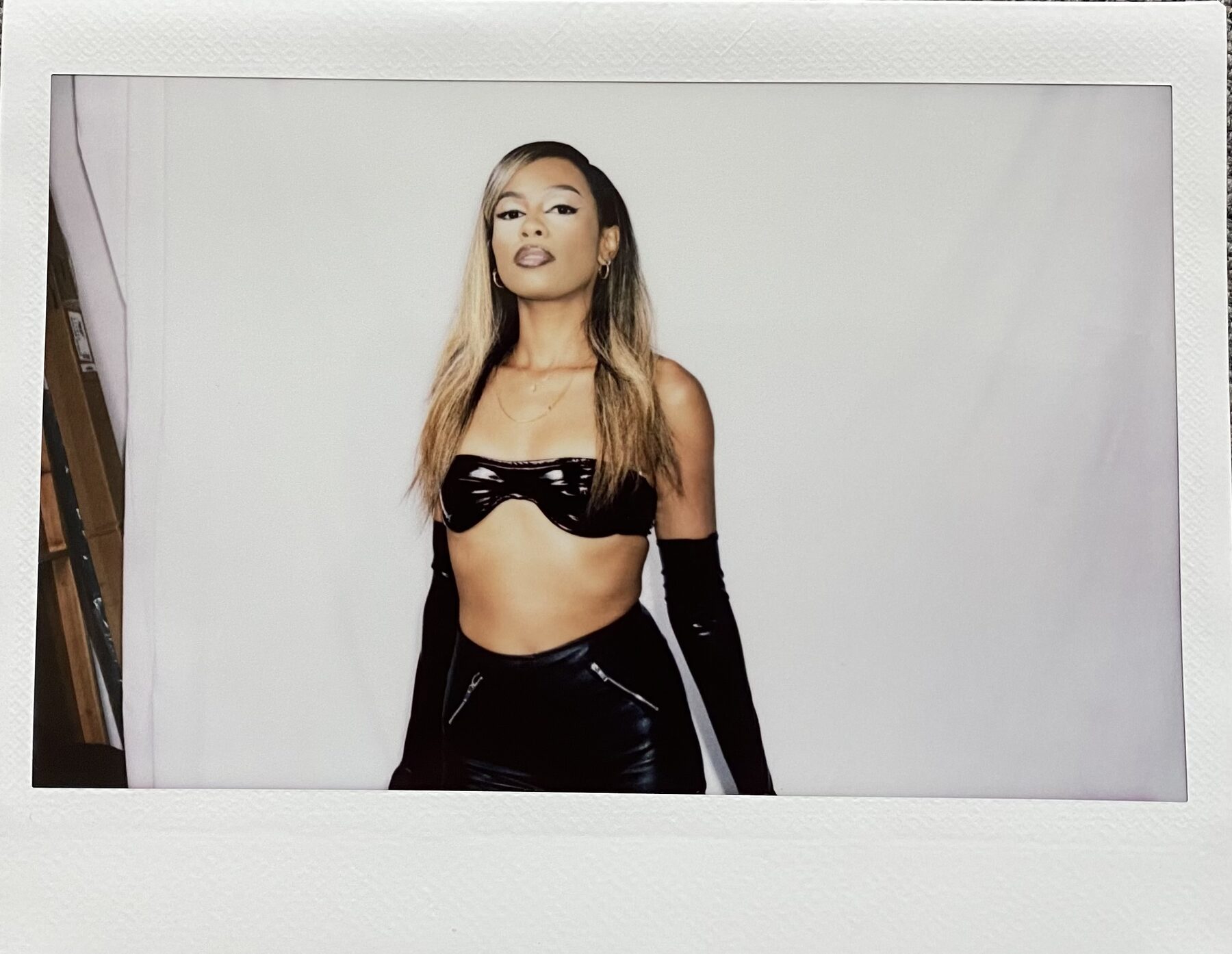 Tash Williamson in an Instax-style photograph against a plain white studio backdrop, standing with a direct, composed gaze; wearing a black patent bandeau top, matching high-waisted patent trousers with silver zip pulls, and long black patent opera gloves, sleek straight hair with blonde balayage falling past her shoulders, a delicate gold necklace at her collarbone.
