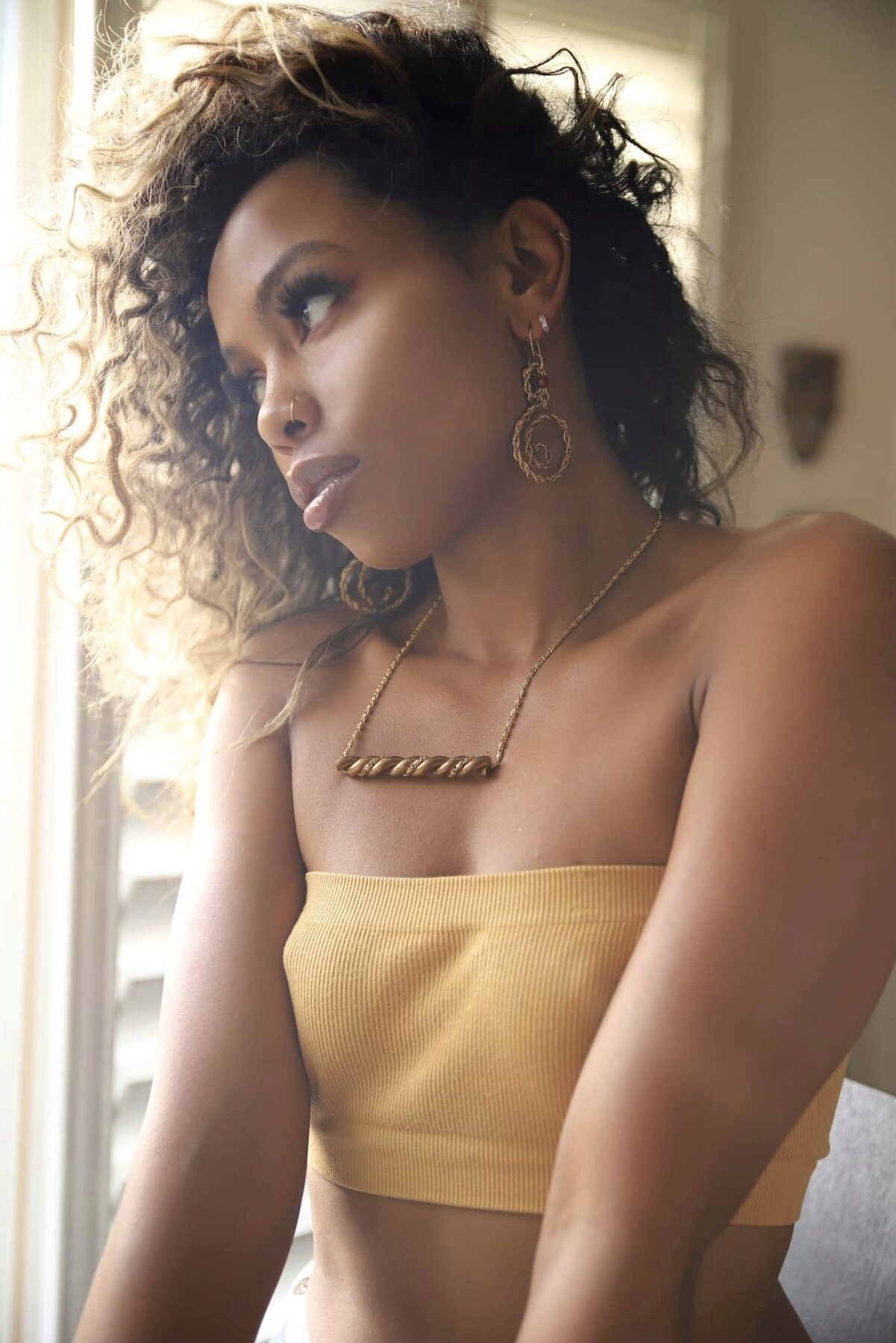 Tash Williamson in a profile beauty shot bathed in warm golden-hour window light, wavy hair swept back loosely with soft tendrils framing her face; bronze smoky eyeshadow and glossy nude lips pair with dangling gold rope-textured spiral earrings and a chunky twisted gold rope necklace, worn with a ribbed mustard tube top as she gazes off to the side.