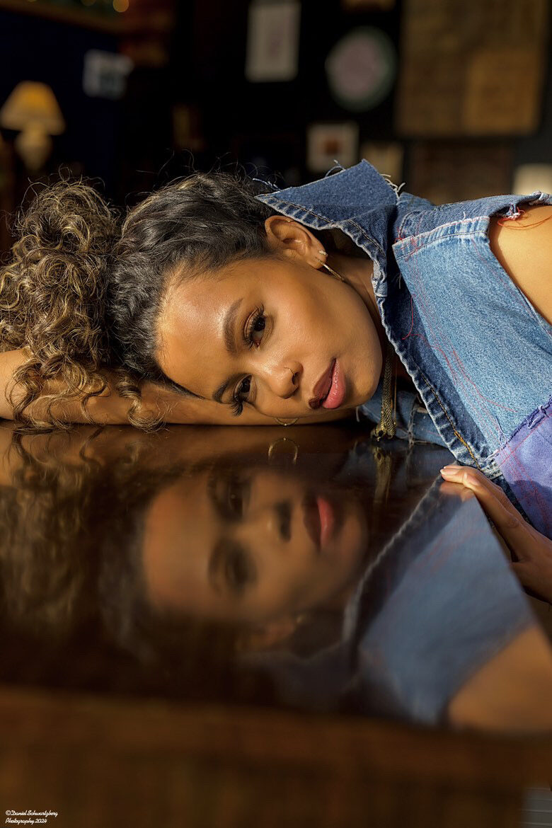 Tash Williamson in a moody close-up lying on her side atop a dark reflective tabletop, her face mirrored in the glossy surface below; curly hair is gathered up loosely with tendrils loose at the temples, softly smoky brown eye makeup and nude glossy lips, a small nose ring and gold hoop earring, wearing a cropped denim jacket over a purple top, low warm lighting with a blurred eclectic background of framed art and a lamp.