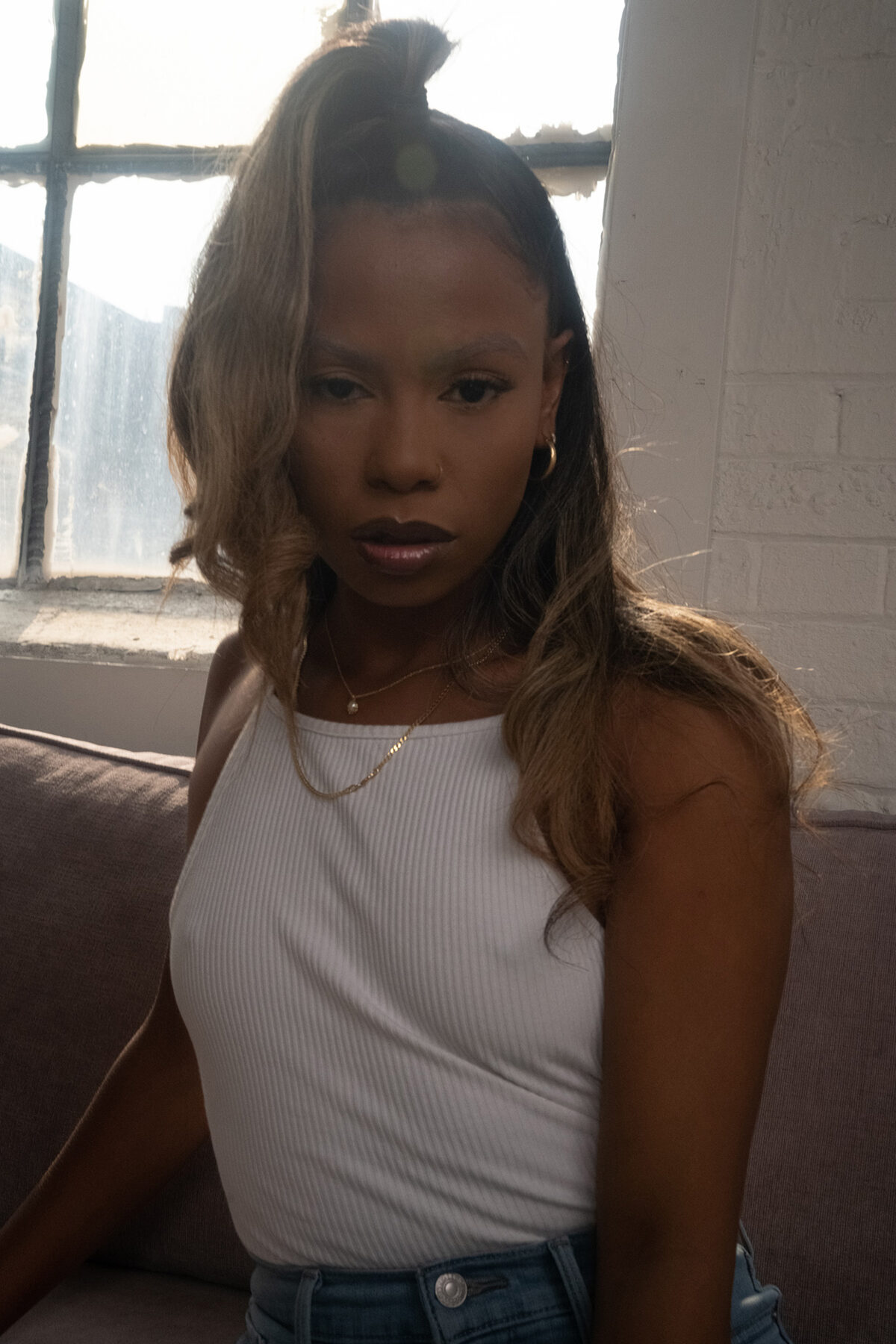 Tash Williamson in a close three-quarter portrait backlit by a tall paned loft window, half-up hairstyle with wavy ash-blonde balayage cascading past the shoulders over dark roots, a sultry heavy-lidded gaze with smoky dark eyeshadow and a deep berry-brown lip, small gold hoop earrings and layered gold necklaces with a tiny pearl pendant, wearing a ribbed white tank top and jeans while seated against a mauve couch in soft window light.