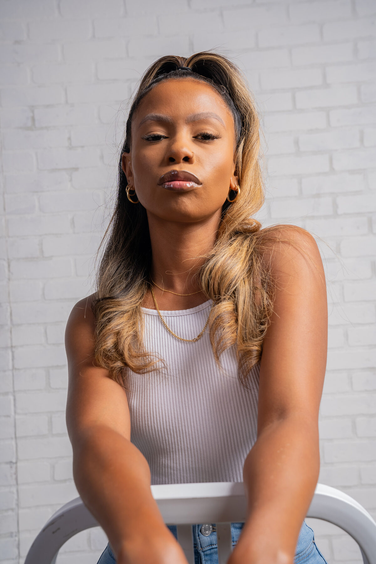 Tash Williamson in a close bust portrait against a white brick studio wall, half-up wavy hair in ash-blonde balayage falling past the shoulders, a direct unsmiling gaze with soft smoky eye makeup and a matte brownish-mauve lip, small gold hoop earrings and a thin gold chain necklace, wearing a ribbed white tank top and seated backward on a white stool with jeans visible; a similar half-up style and tank top to a companion loft-window shot, but here set against a plain brick backdrop with flatter studio light and a more square-on pose.