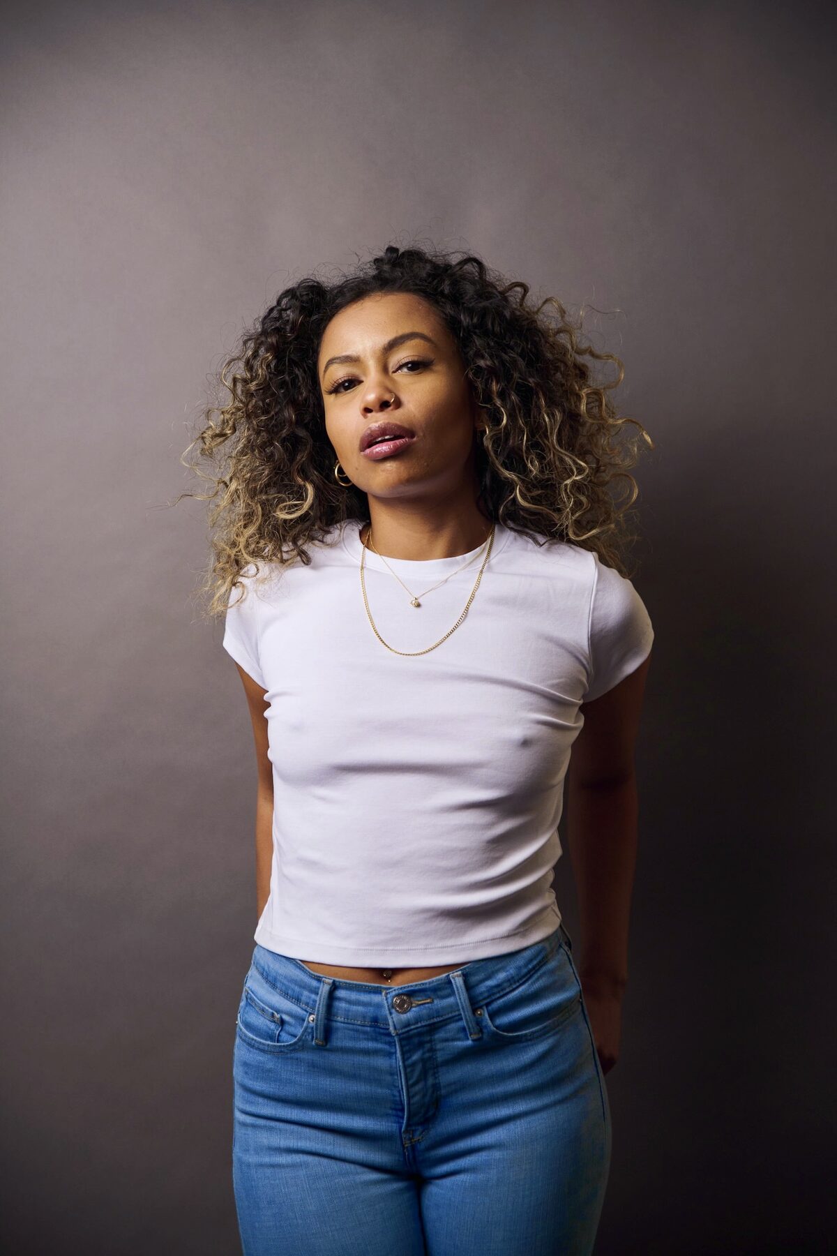 Tash Williamson in a moody studio headshot against a plain charcoal-grey backdrop, full curly hair in a blonde-and-brunette balayage worn loose and voluminous around the face, a direct steady gaze with soft smoky eye makeup, a nose ring, and a glossy mauve-brown lip, delicate layered gold necklaces with a small pendant over a plain white crew-neck tee, dramatic side lighting casting soft shadow across one side of her face.