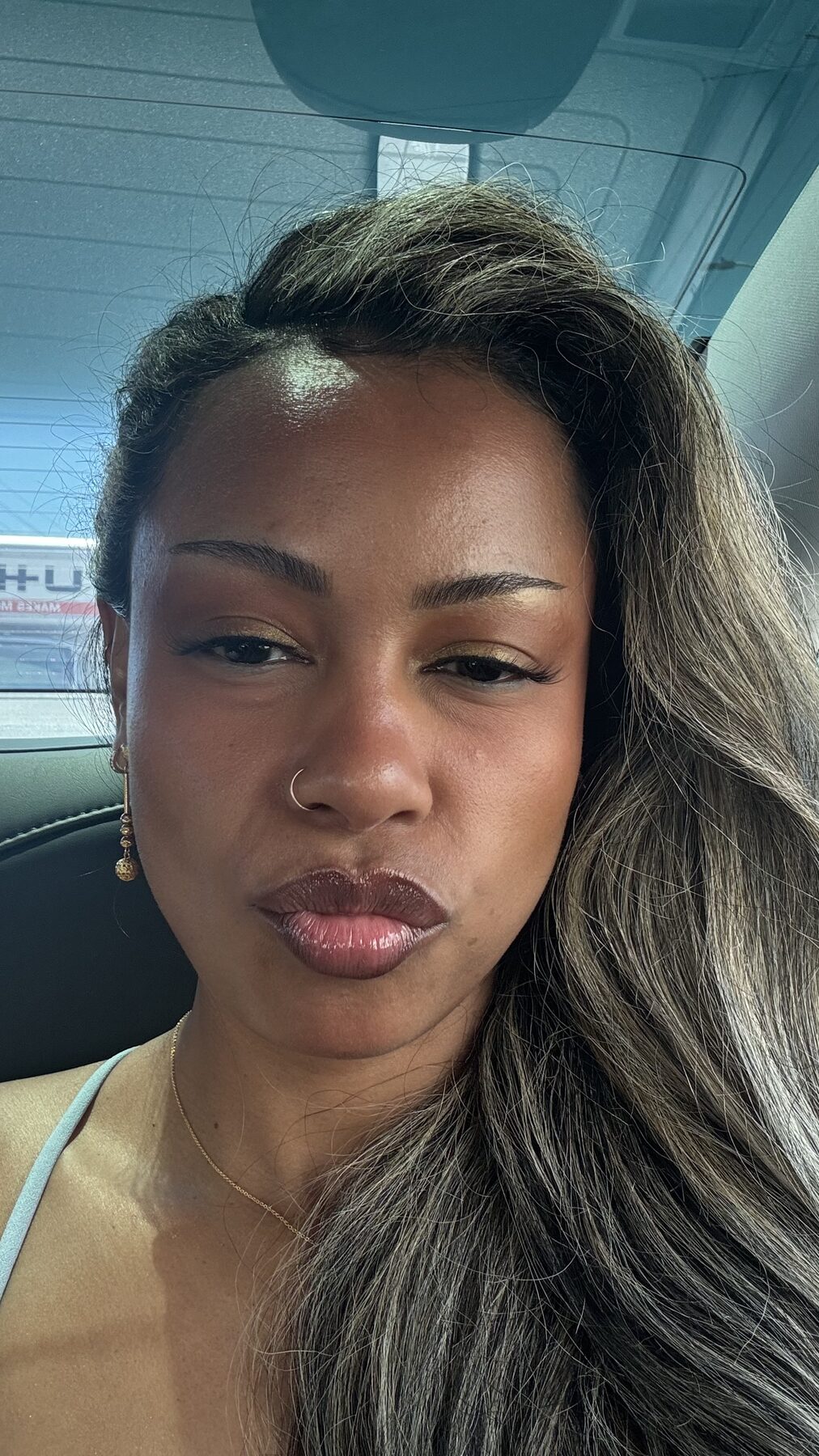 Tash Williamson in a close-up car selfie, chin tilted down and lips pursed in a soft pout, long wavy hair with grey-blonde balayage falling loose over one shoulder, warm golden eyeshadow with defined winged liner and a glossy brown-mauve lip, a nose ring, a dangling beaded drop earring, and a fine gold necklace, wearing a pale blue halter top in natural daylight through the car window.