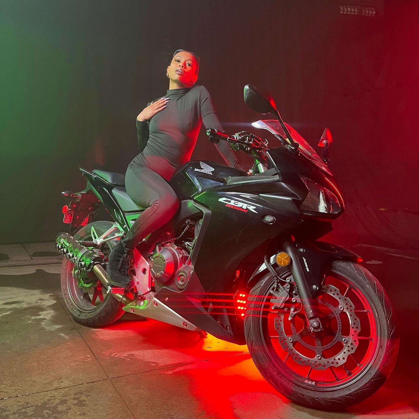 Tash Williamson seated sideways on a black Honda CBR500R sport motorcycle in a dark studio bay lit with dramatic red and green gels, one hand pressed flat to her chest, chin lifted with a direct, sultry gaze into the camera; wearing a fitted dark olive long-sleeve turtleneck bodysuit and black boots resting on the bike's foot pegs, hair slicked back, small hoop earrings.