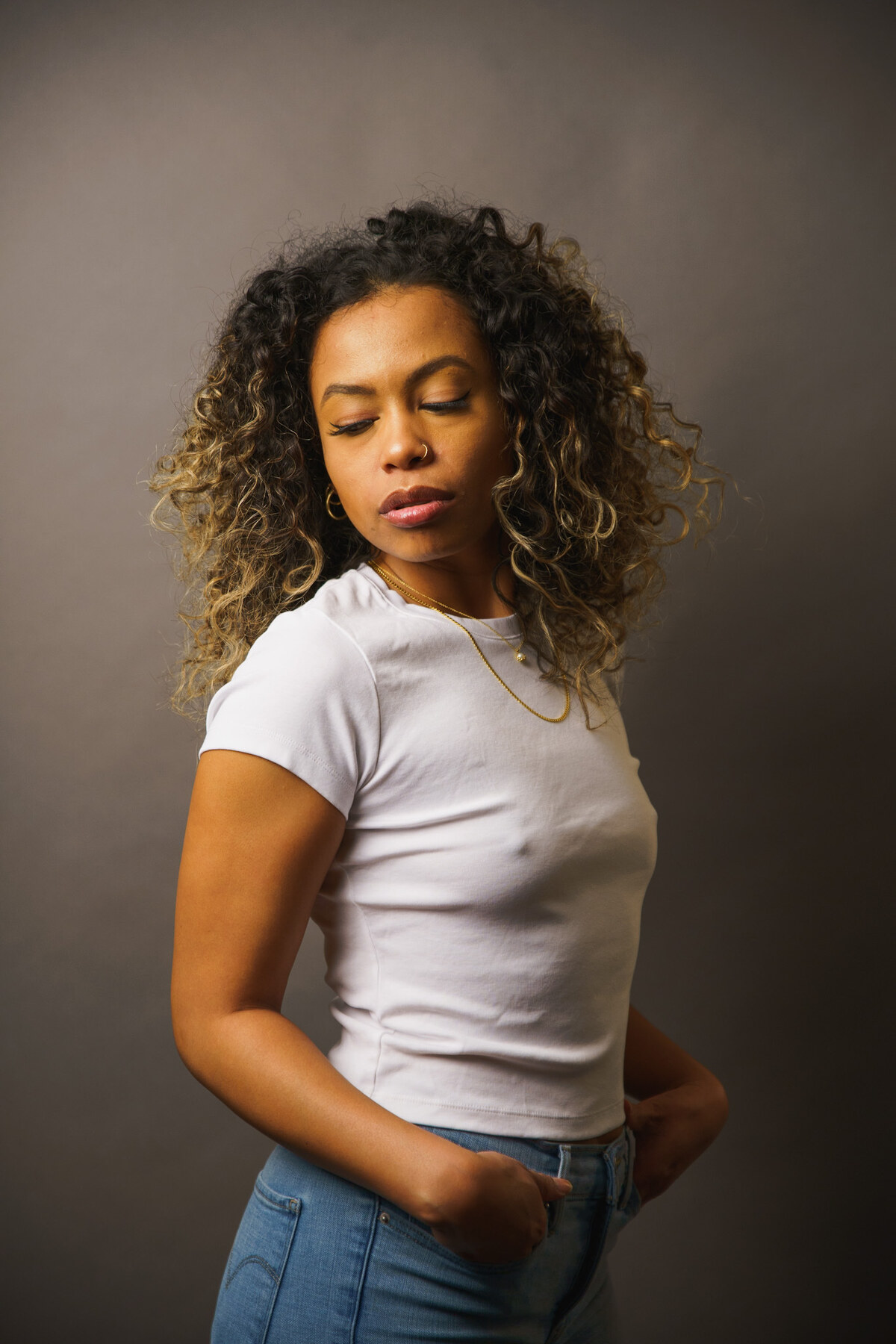 Tash Williamson in a three-quarter studio portrait against a plain warm-gray backdrop, chin dipped and eyes cast down with a calm, closed-mouth expression, dramatic side lighting catching her face and voluminous curly balayage hair; wearing a fitted white crop tee and blue jeans with both hands tucked into her back pockets, a small gold nose ring and layered fine gold necklaces at her throat.