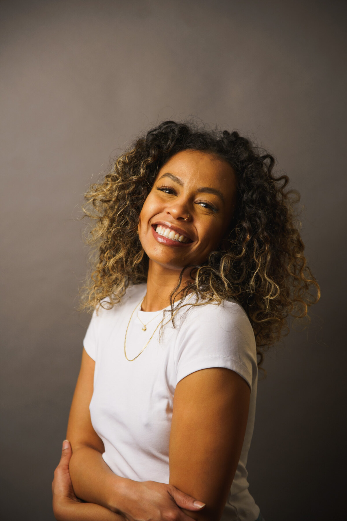 Tash Williamson in a warm, backlit studio headshot from the same white-tee session as two companion shots, head tilted with arms crossed, laughing with a wide open smile and bright teeth; the same voluminous curly balayage hair and layered gold necklaces, golden rim light outlining her hair against the plain backdrop.