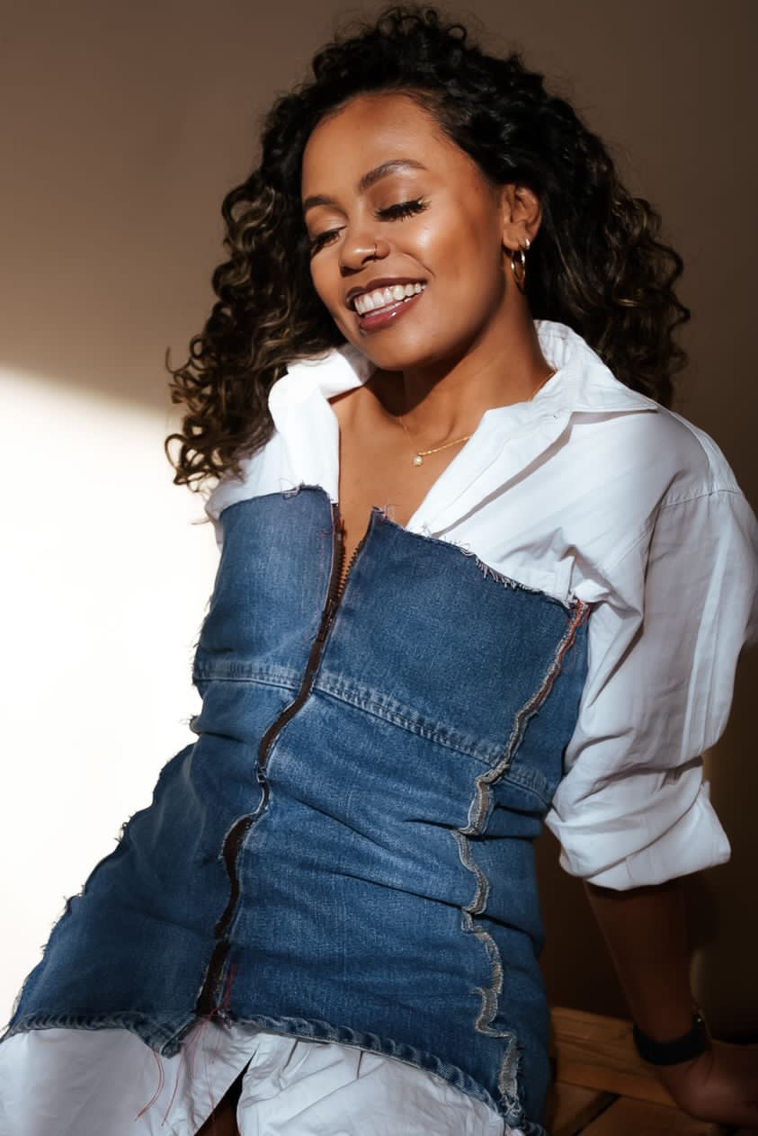 Tash Williamson laughing brightly with her eyes closed and head tilted down, warm directional sunlight raking across her face; wearing a white collared button-down layered under a dark denim zip-front pinafore dress with raw frayed seams, curly dark hair with soft highlights, set against a plain sunlit wall with a wooden ledge at the edge of the frame.