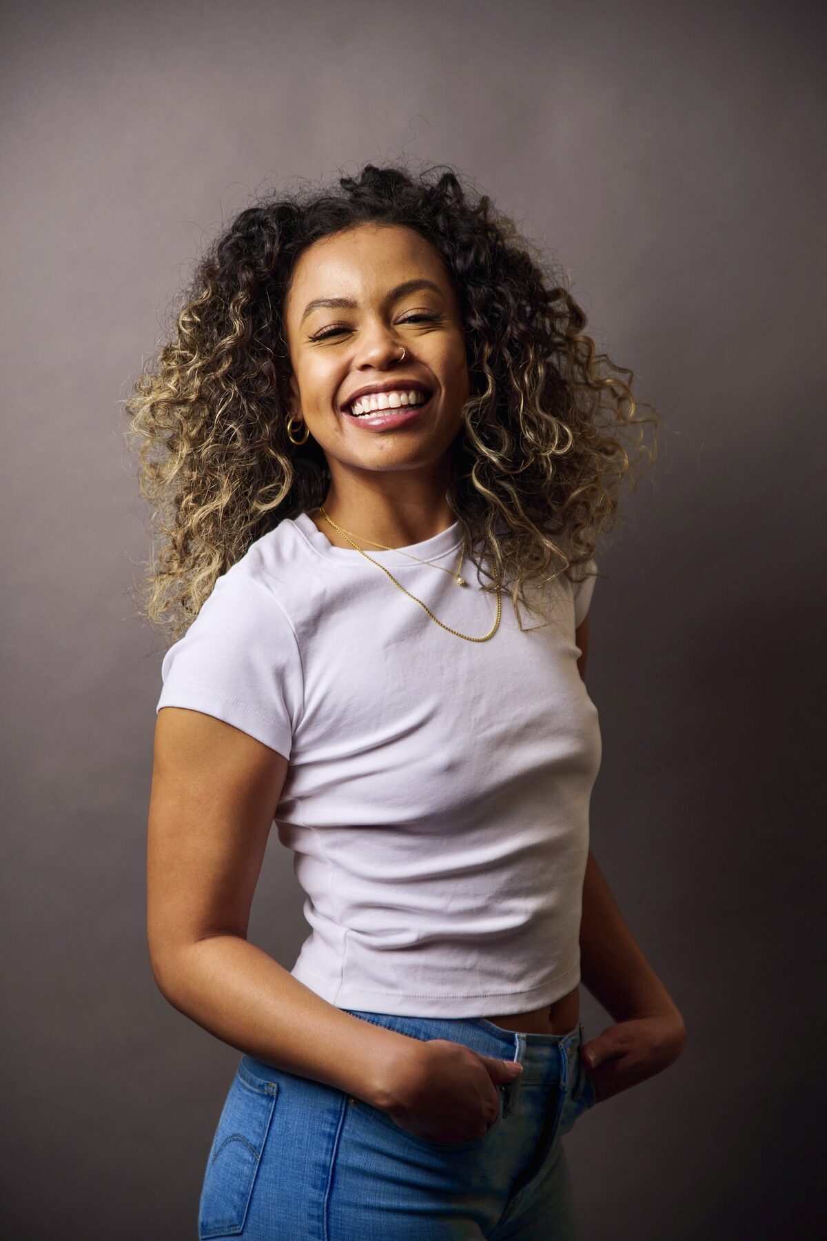 Tash Williamson in a three-quarter studio portrait from the same white-tee session as two companion shots, laughing with her head tilted and eyes closed in a genuine, joyful expression, one hand tucked in her jeans pocket, curly balayage hair loosely tousled, warm-gray backdrop and soft directional light.