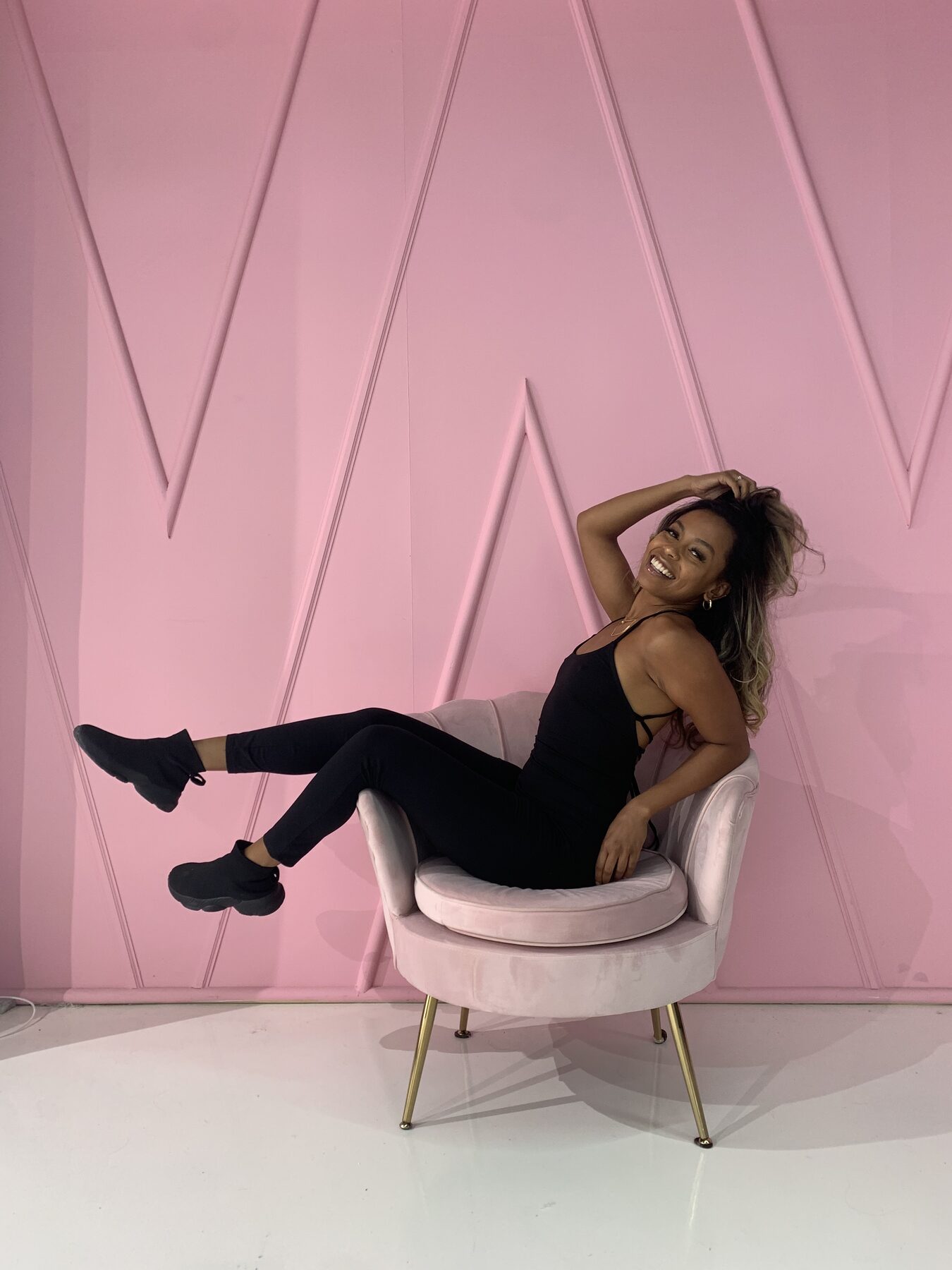 Tash Williamson caught mid-motion, one hand tousling her wind-blown hair as she leans back over the arm of a blush-pink velvet chair with a bright open smile, wearing a black strappy bodysuit, black leggings, and black platform sneakers, set against a glossy pink paneled studio wall — a playful, energetic action shot rather than a posed portrait.