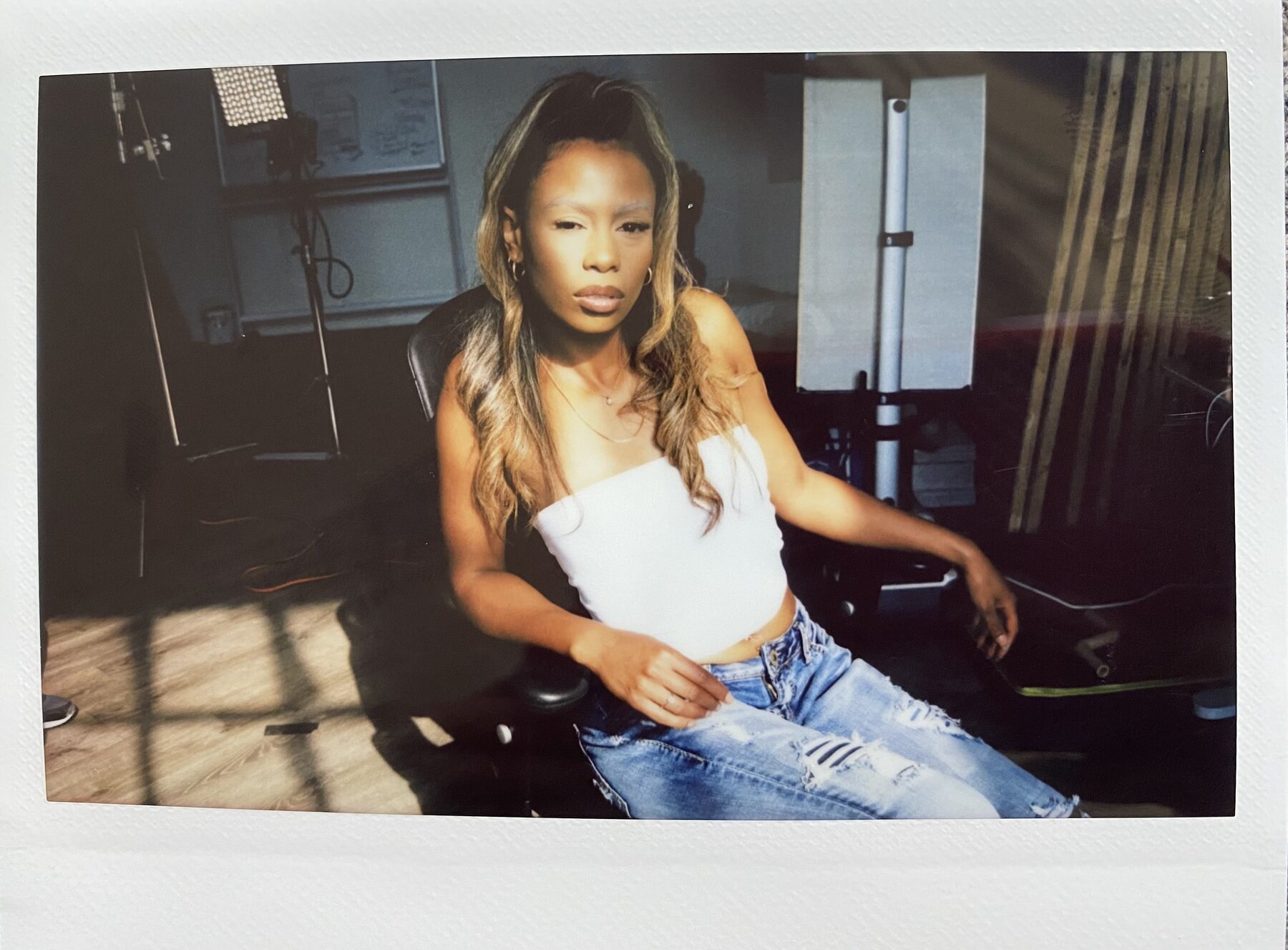 Tash Williamson in a candid Polaroid-style behind-the-scenes shot, seated in a black studio chair with one leg crossed, a calm, composed expression looking directly at the camera; wearing a white strapless top and distressed blue jeans, wavy hair loose past the shoulders, studio lighting equipment, cords, and a whiteboard visible in the background.