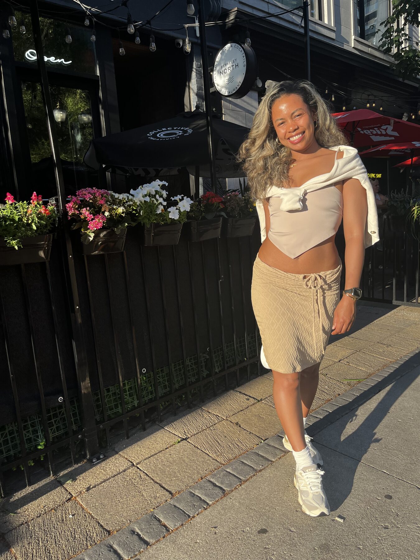 Tash Williamson smiling brightly outdoors on a restaurant patio, walking with one arm swinging and a gold watch visible on her wrist, wearing a white halter top tied at the waist over a tan textured mini skirt and white sneakers, wavy hair loose, string lights and flowering planters framing the entrance behind her with a round sign reading "North" on the building.
