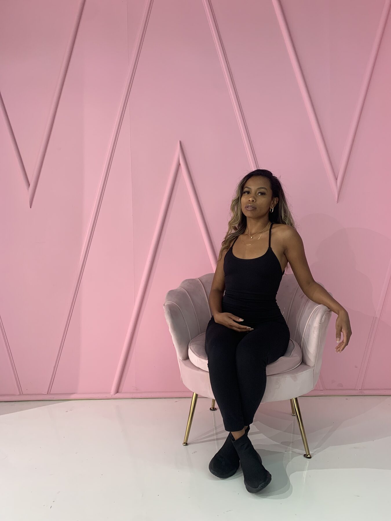 Tash Williamson seated in the same blush-pink velvet chair and glossy pink paneled studio as a companion shot, one arm raised gracefully overhead, gazing calmly toward the camera; wearing a black scoop-neck cami top, black leggings, and black ankle boots, a thin gold necklace at her throat.