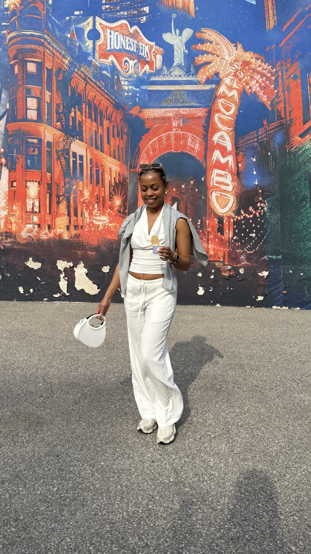 Tash Williamson walking and smiling outdoors past a colorful mural of a vintage cityscape, holding a wafer-cone ice cream in one hand and a white top-handle bag in the other, a light gray cardigan slipping off her shoulders over a white wrap top and cream drawstring trousers, sunglasses pushed up into her hair, white sneakers.