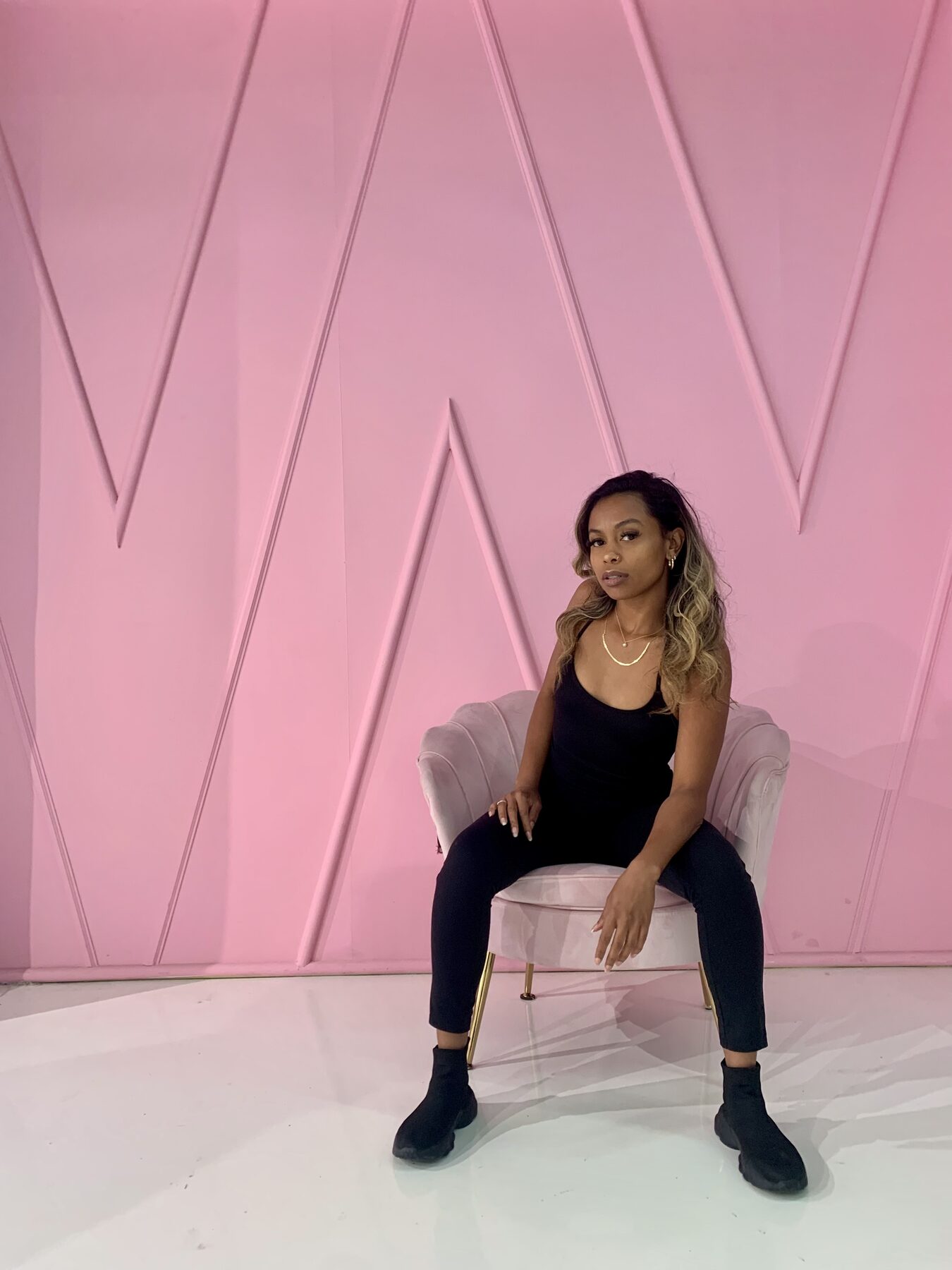Tash Williamson seated upright in the same blush-pink velvet chair from the pink-paneled studio set, legs crossed at the knee with black ankle boots, one hand resting on her knee, meeting the camera with a steady, composed gaze; wearing a black scoop-neck bodysuit, hair loose in soft waves with a gold hoop earring visible.
