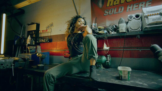 Tash Williamson seated on a cluttered metal workbench inside an auto garage, one leg drawn up and the other braced against the table edge, one hand raised near her chin in a pensive, camera-aware expression; wearing a black tank top and olive cargo-style pants, loose curly hair, tools, parts bins, and a red-and-white "Havoline Sold Here" sign visible on the wall behind her — a cinematic, film-still mood rather than a straight studio shot.