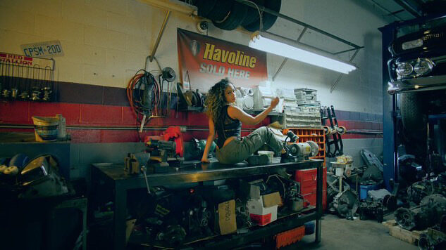 Tash Williamson in the same auto-garage workbench set as a companion shot, caught mid-motion twisting toward a wall-mounted shelf of tools and parts; same black tank top, olive cargo pants, and loose curly hair, workbench clutter and the "Havoline Sold Here" sign in the background — an alternate angle from the same cinematic sequence.