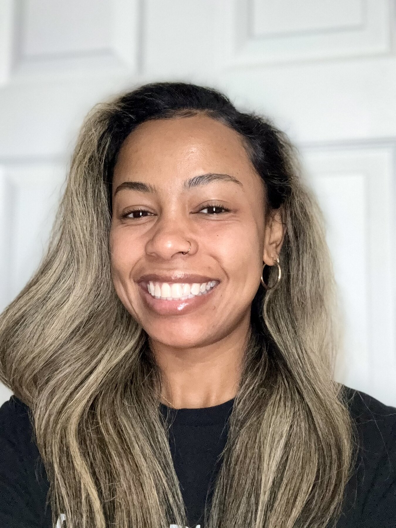 Tash Williamson smiling broadly at the camera in a straight-on headshot against a plain white paneled door, wavy hair with ash-blonde balayage over dark roots worn loose past the shoulders.