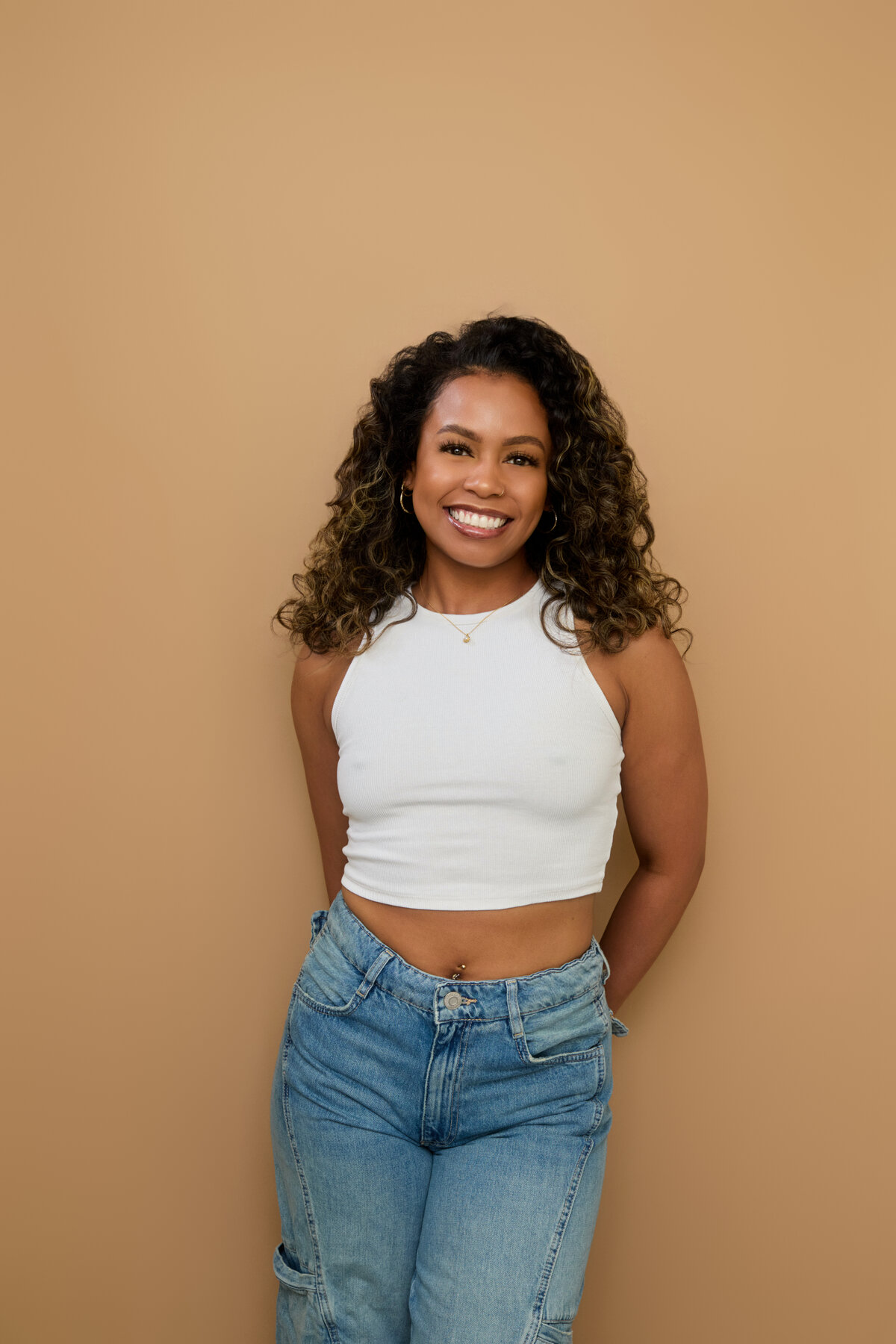 Tash Williamson smiling with teeth showing in a waist-up studio portrait against a warm tan backdrop, hands clasped behind her back, wearing a white halter crop top and light-wash jeans, curly hair with blonde highlights worn loose.
