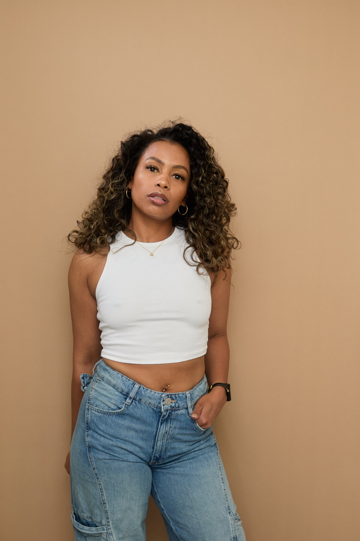 Tash Williamson standing in a waist-up studio portrait against a warm tan backdrop, one hand tucked in her jeans pocket, wearing a white halter crop top, curly hair loose, expression calm and serious.