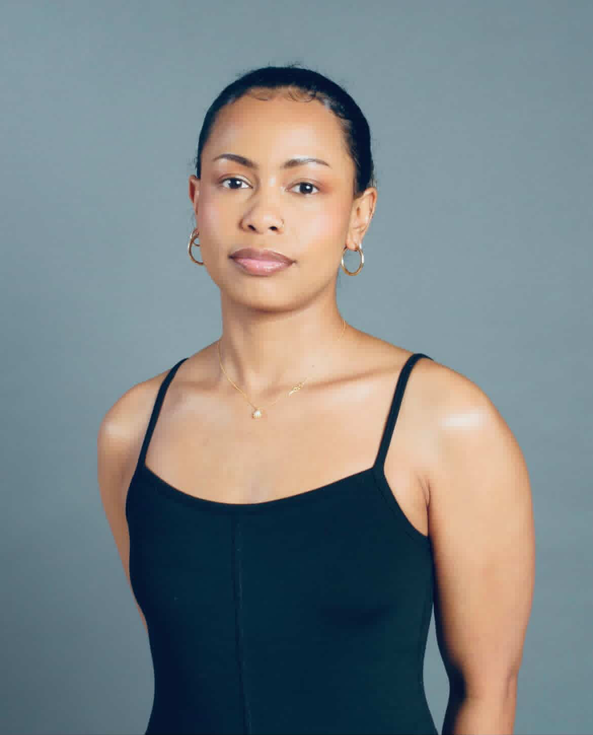 Tash Williamson in a studio headshot against a plain blue-gray backdrop, hair pulled back sleek off the face, wearing a black cami top, with a soft, direct gaze.