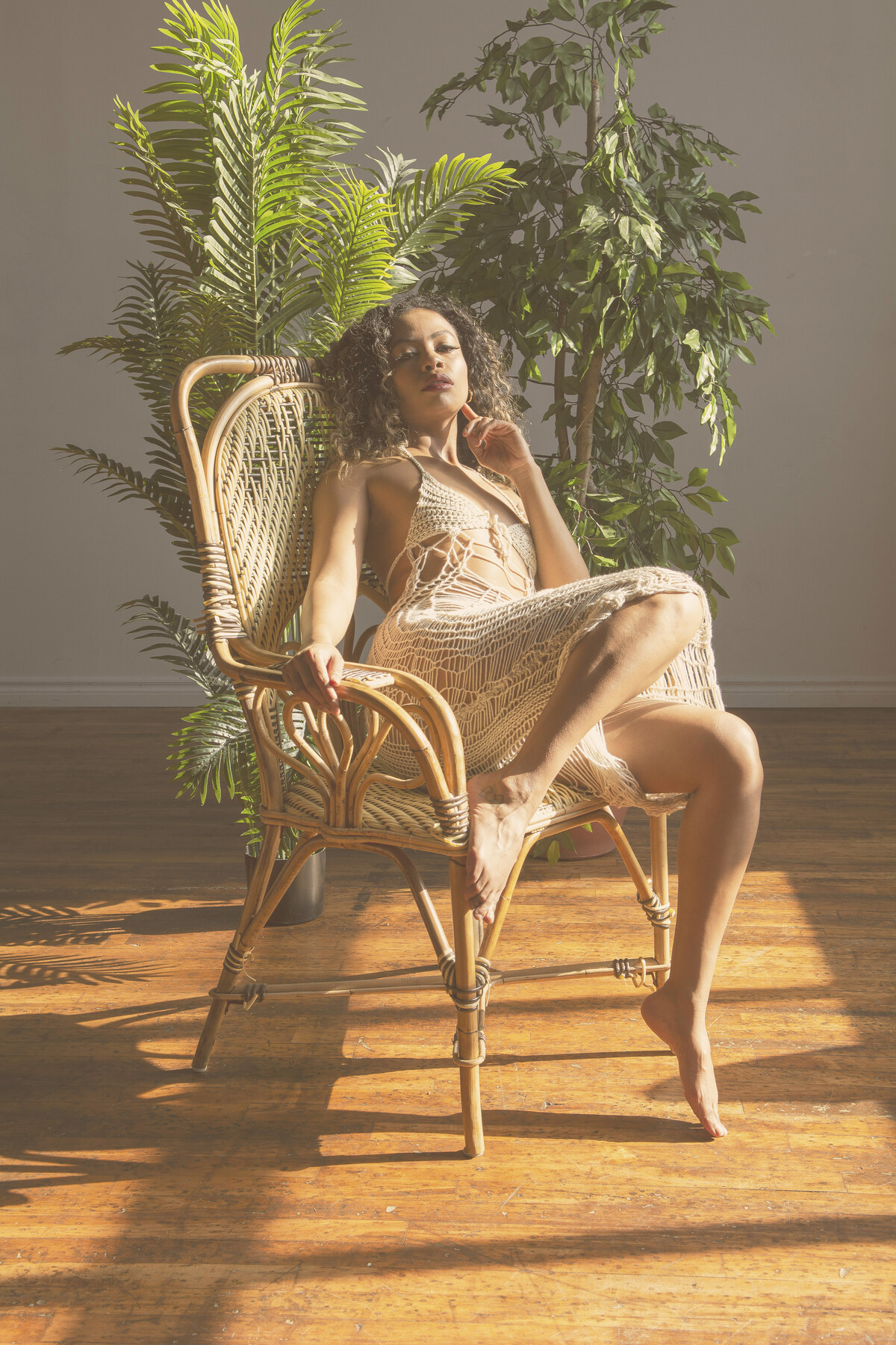 Tash Williamson in a full-length seated portrait in a rattan wingback chair, wearing a sheer cream crochet dress, one leg bent up onto the chair seat and the other extended to the floor, one hand resting near her chin and the other on the armrest, wavy dark curls with blonde balayage, gazing directly at the camera with sunlit potted palms behind her.