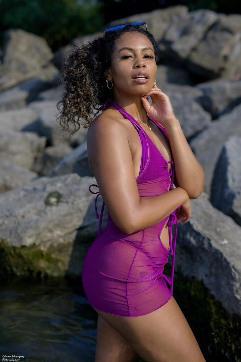 Tash Williamson in an outdoor portrait standing among large lakeside boulders, wearing a sheer magenta halter mini dress with tie-side straps, sunglasses pushed up into her curly ponytail, one hand raised to her jaw, lips parted as she looks back toward the camera.