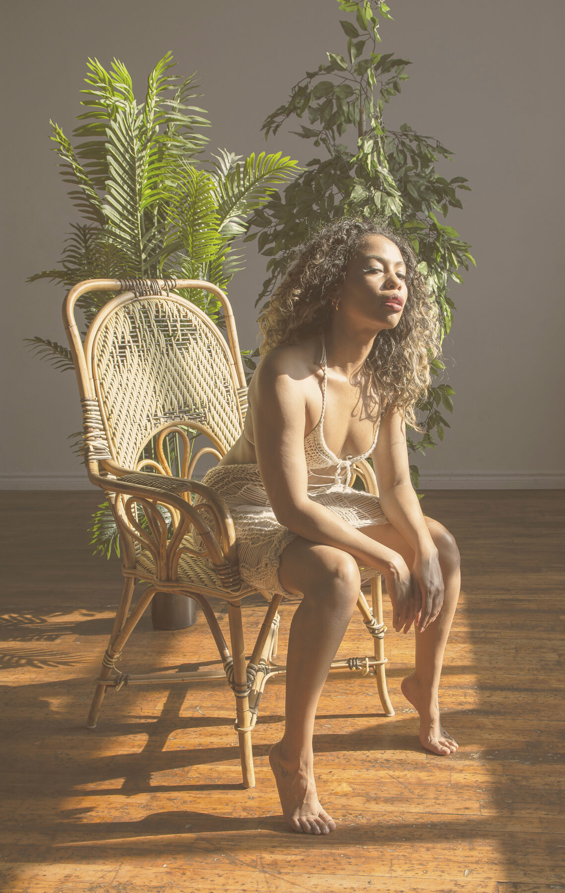 Tash Williamson in the same rattan chair and crochet dress as a companion shot, this time reclined back with head tilted and eyes softly closed, one hand lightly touching her curls near her shoulder and legs crossed at the knee, hazy backlit sun filtering through the plants behind her.