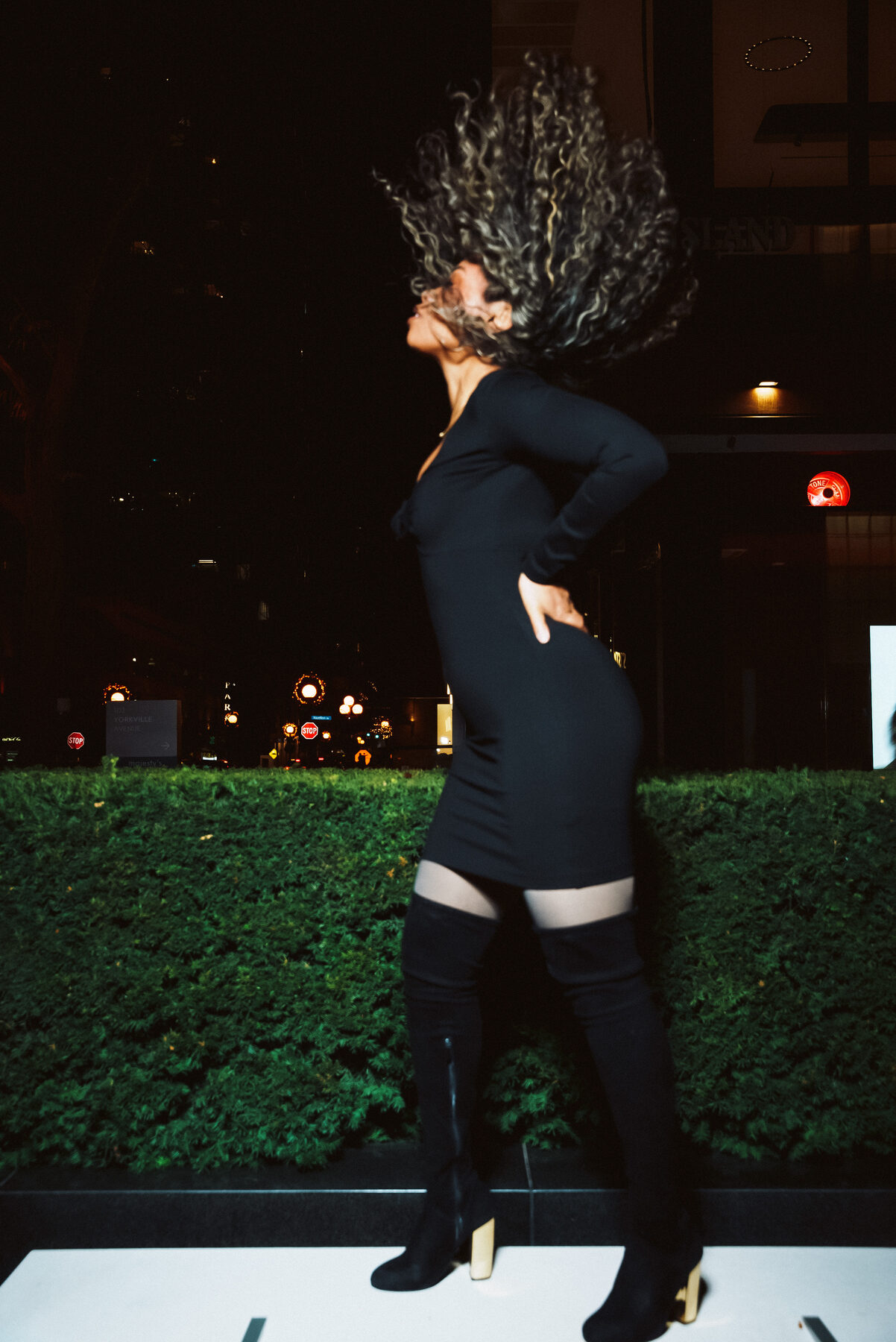 Tash Williamson in a full-length night street shot, caught mid hair-toss with her curly hair flying upward, one hand on her hip in profile, wearing a fitted black long-sleeve mini dress and sheer-paneled thigh-high boots, standing before a hedge with a lit street sign and city lights behind her.