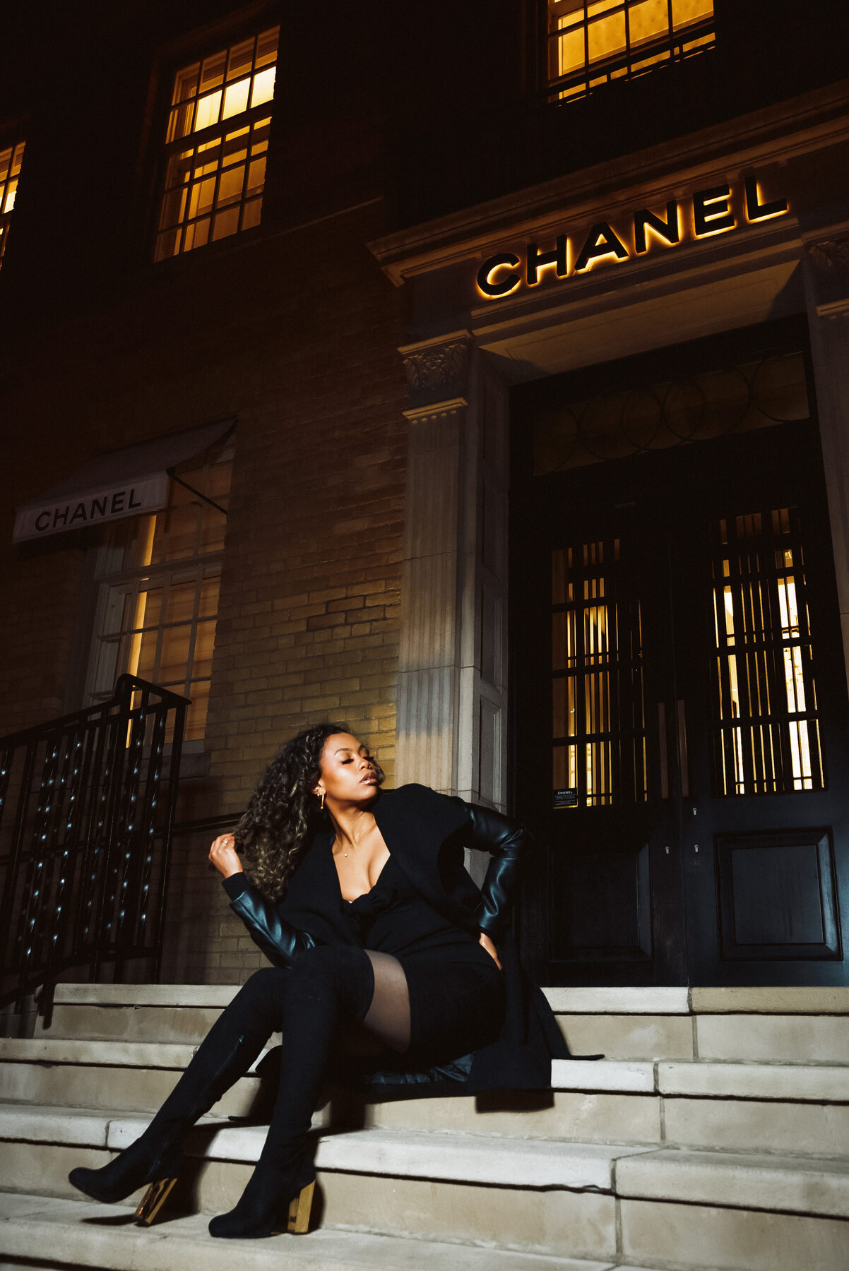 Tash Williamson in a full-length low-angle shot seated on the stone steps outside a lit Chanel storefront at night, leaning back on one braced arm with the other hand resting on her hip, legs crossed at the knee, chin dipped and eyes closed in an unguarded moment, wearing an open black wrap coat with leather sleeves over a black mini dress and thigh-high suede boots with gold block heels, curly hair loose over one shoulder.