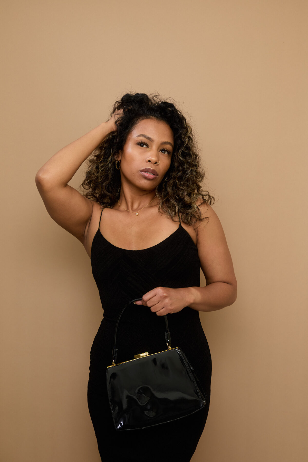 Tash Williamson in a studio waist-up portrait against a warm beige backdrop, one hand raised into her curly hair and the other holding a black patent vintage box purse with a gold clasp at her side, black textured spaghetti-strap slip dress, gold necklace and nose ring, gazing off to the side with a serious, sultry expression.