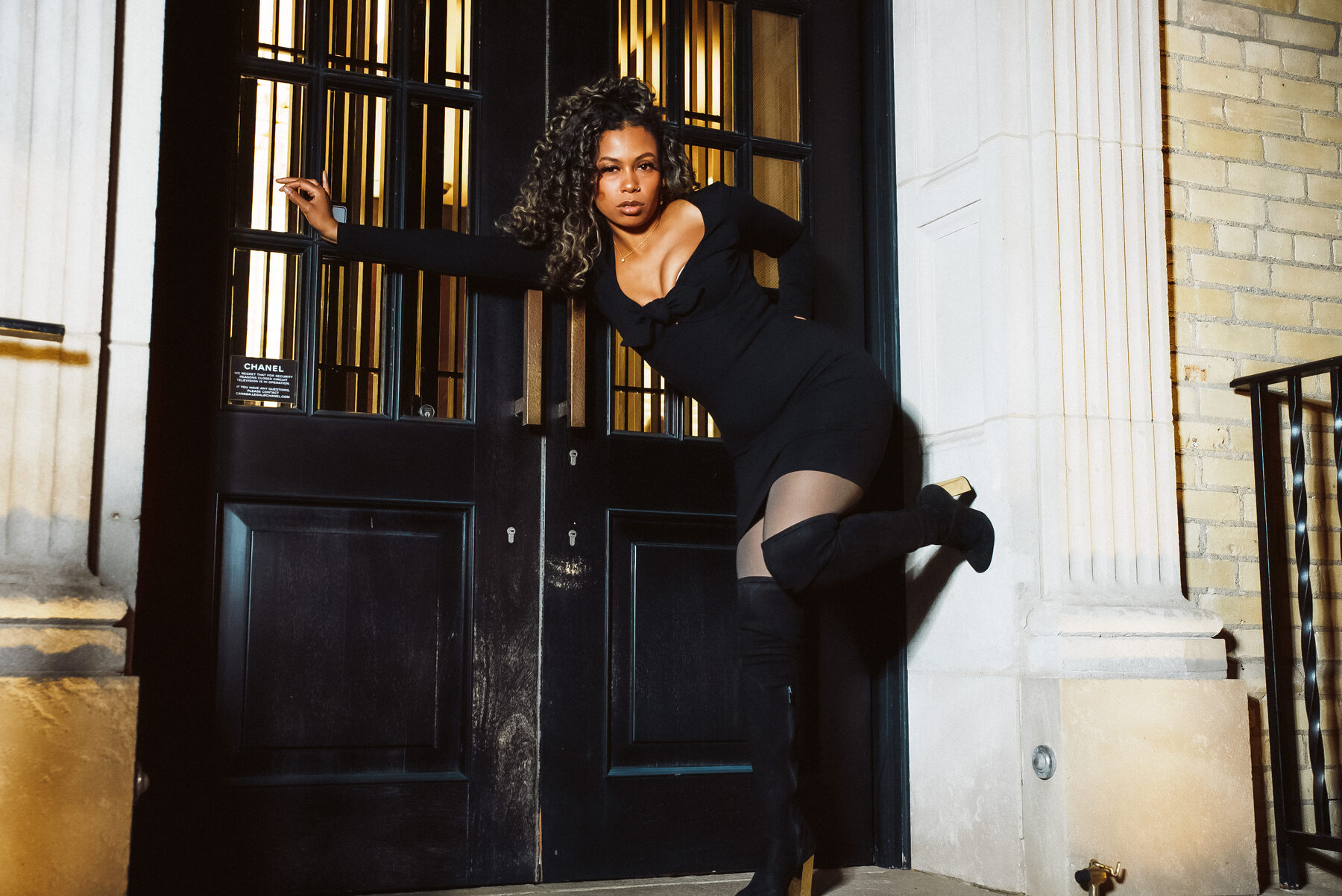 Tash Williamson in a close, dynamic frame leaning sideways into the Chanel doorway at night, one arm stretched overhead gripping the door's iron grille and one knee bent and lifted against the frame, a direct sultry gaze at the camera, off-shoulder black mini dress with a plunging neckline, sheer tights and thigh-high boots, curly hair falling loose.
