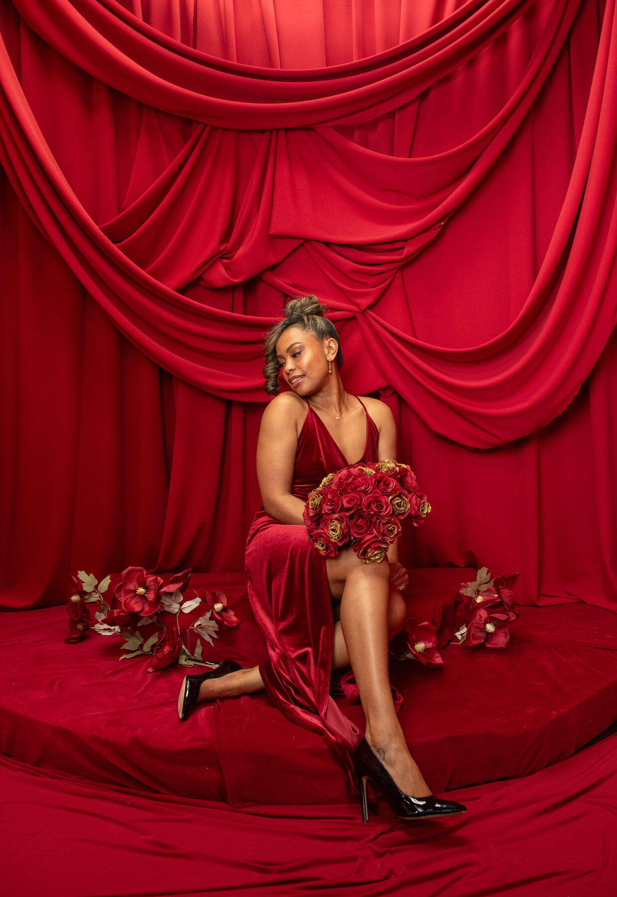 Tash Williamson kneeling on a red-draped stage, legs folded to one side, a red velvet halter gown with a high slit, a bouquet of red and gold roses cradled in her lap, hair swept into a bun with curled tendrils framing her face, looking down and to the side with a soft smile, black pointed pumps, faux flowers scattered on the platform around her.