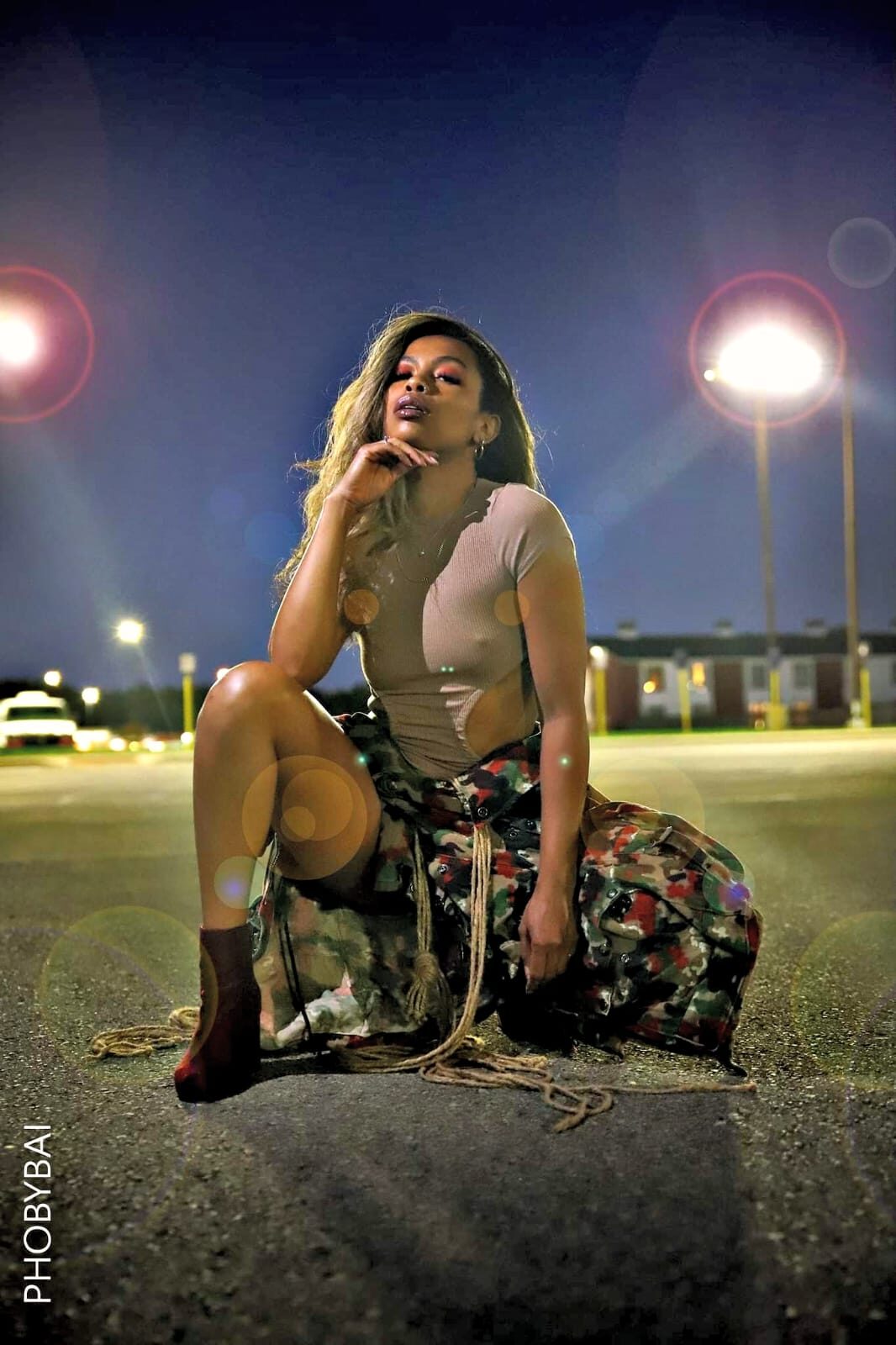 Tash Williamson crouched low in a nighttime parking lot, wearing a nude short-sleeve top and a fringed camo maxi skirt with cargo pocket detailing, one hand raised near her chin, dark ankle boots, waves of blonde-highlighted hair loose around her shoulders, dramatic lens flare from streetlights glowing behind her.