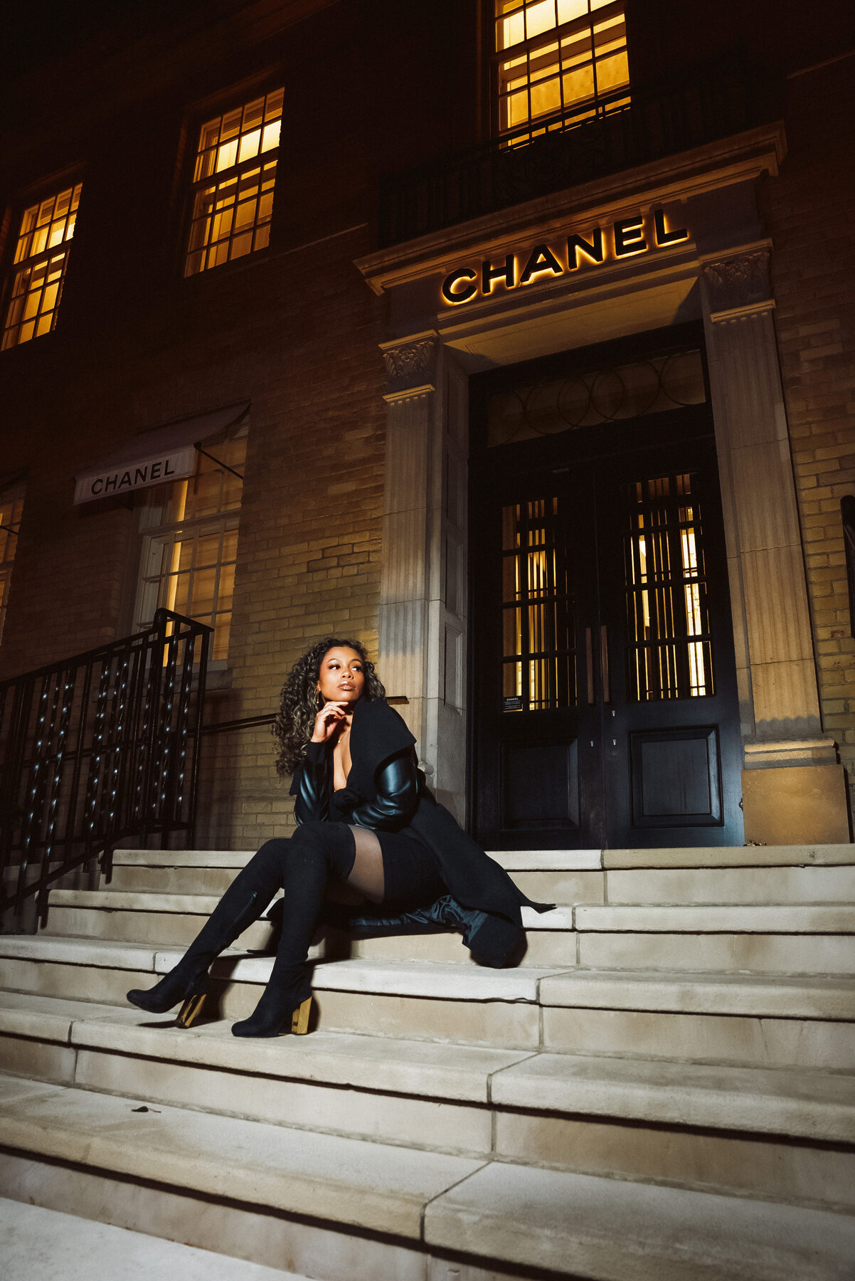 Tash Williamson in a full-length shot seated on the storefront steps at night, one hand raised thoughtfully to her cheek as she looks off to the side in near-profile, the other arm relaxed at her side, black leather-sleeved coat over a mini dress, thigh-high boots crossed at the ankle, curly hair with blonde highlights falling forward.