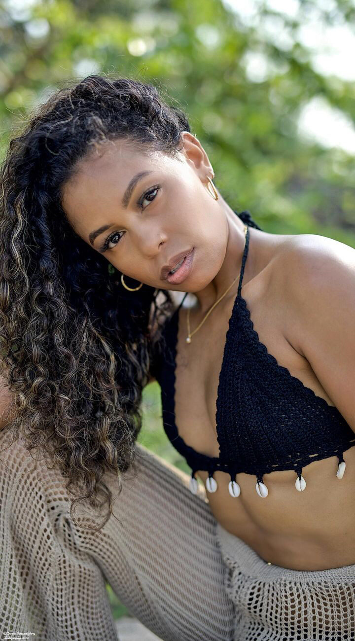 Tash Williamson in an outdoor close-up portrait framing her head and chest, leaning in close to the camera, a black crochet halter top trimmed with cowrie shells, wavy curly hair with blonde highlights cascading over one shoulder, soft greenery blurred behind her, gold jewelry, a direct, sultry gaze.