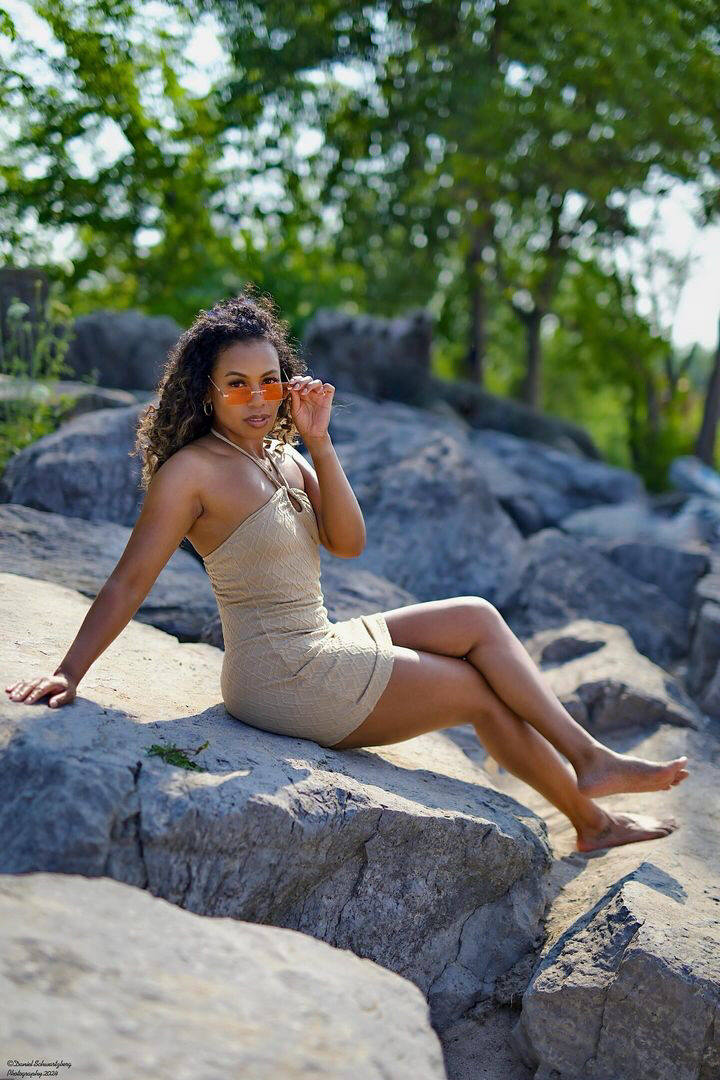 Tash Williamson in a full-length seated portrait on lakeside rocks, wearing a fitted beige textured halter mini dress, sunglasses lowered partway down her nose with one hand touching them and the other braced on the rock behind her, legs crossed at the knee, bare feet, loose curly hair, wooded greenery blurred behind her.