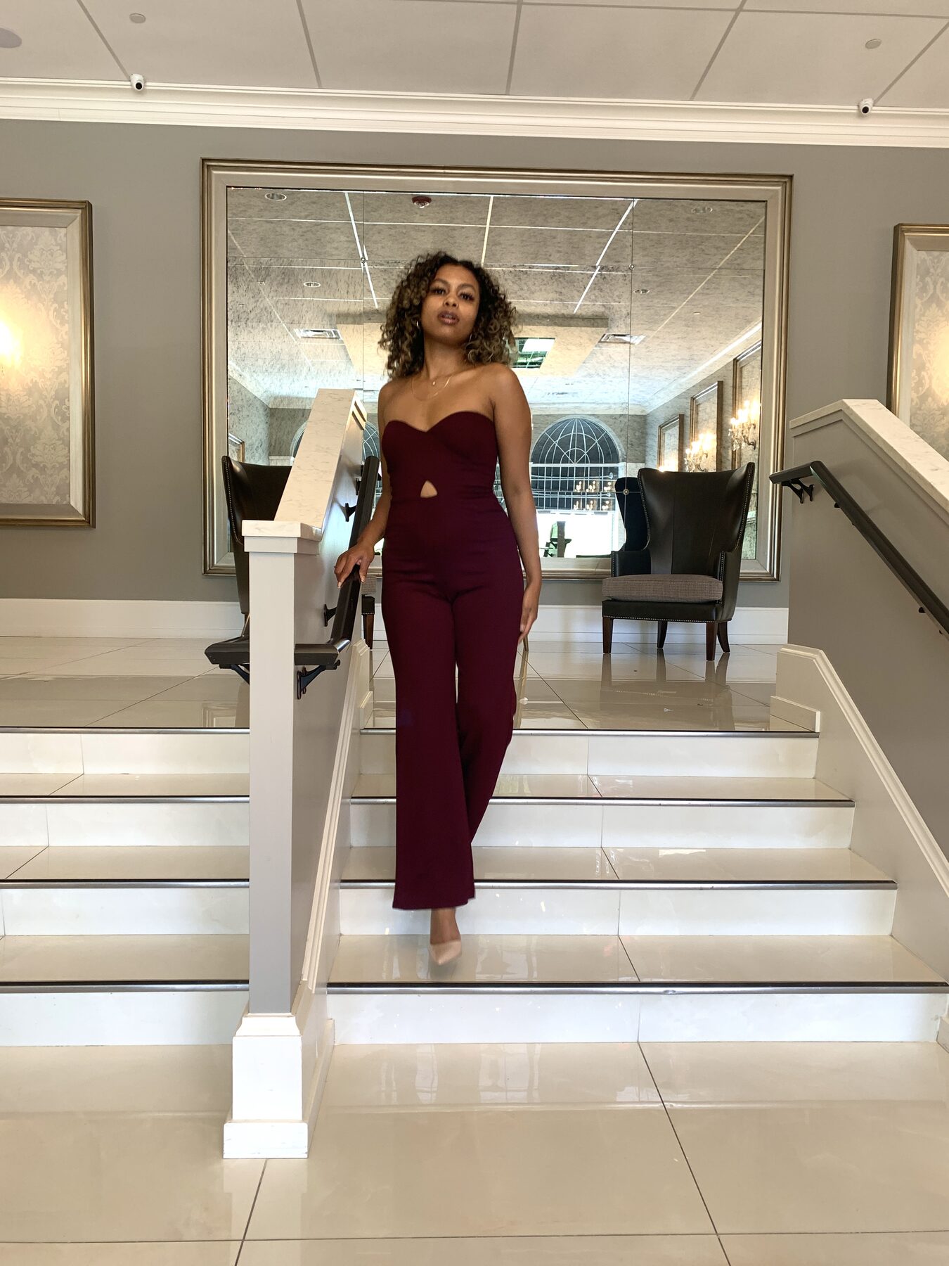 Tash Williamson descending a curved staircase in an elegant hotel lobby, one hand resting on the marble banister, wearing a strapless burgundy jumpsuit with a keyhole cutout at the waist and wide-leg trousers, nude pointed pumps, soft curly hair and a direct gaze toward the camera, mirrors, wingback chairs, and a chandelier reflected in the background.