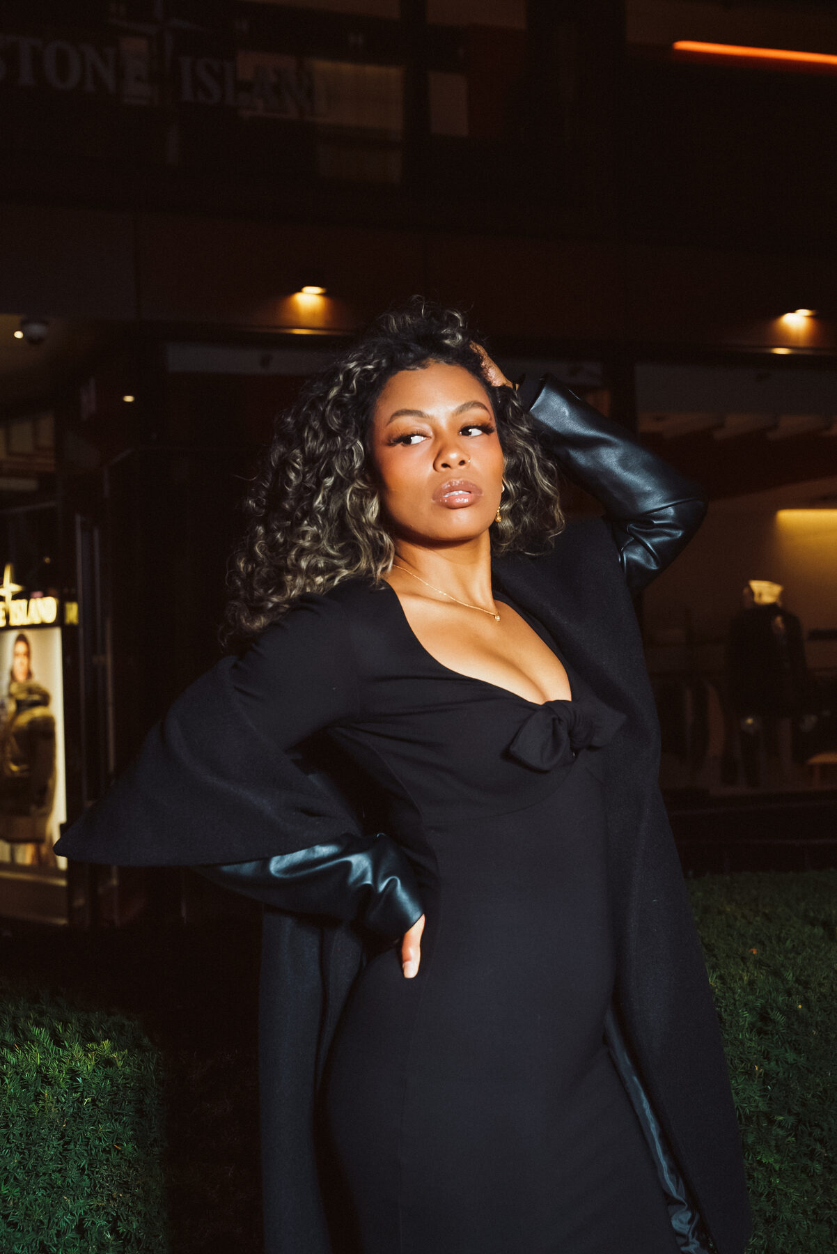 Tash Williamson in a waist-up night portrait, one hand raised into voluminous curly balayage hair as she gazes off to the side, wearing a black plunge-neck dress with a bow at the bust and glossy black pleather sleeves, backlit by an illuminated storefront sign on a city street.