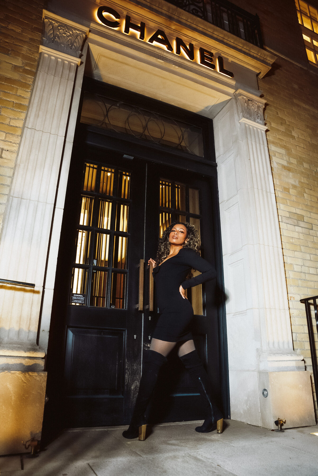 Tash Williamson in a full-length shot standing at the Chanel entrance, one arm raised straight overhead against the door frame and the other hand on her hip, legs crossed at the ankle with her weight shifted into one hip, a confident half-smile toward the camera, black mini dress with the open coat trailing behind her, thigh-high boots.