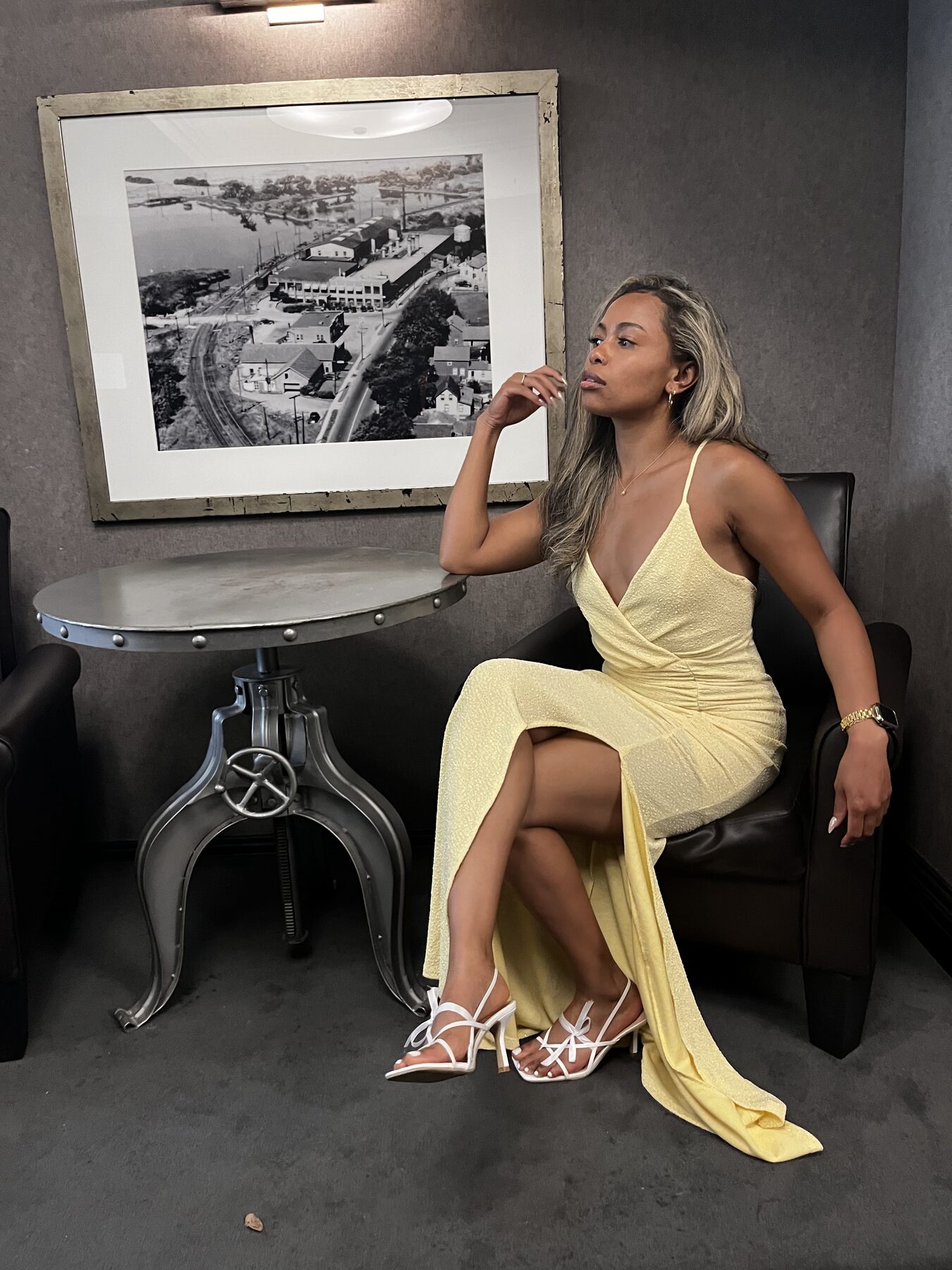 Tash Williamson in a full-length reclining pose on a gray lounge chair, one arm raised overhead resting on the seatback, wearing a pale yellow textured slip dress with a draped cowl skirt and white strappy heels, legs crossed at the ankle, a framed black-and-white photograph propped against the wall beside her.