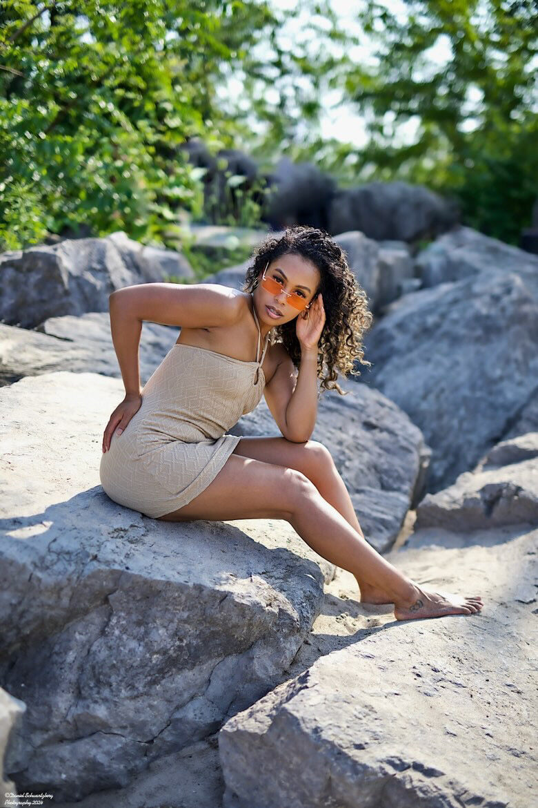 Tash Williamson seated on a rocky outcrop in a tan ribbed bodycon dress, wearing tinted orange sunglasses, one hand resting near her temple and hair and the other braced behind her on the rock, legs crossed with an ankle tattoo visible, soft-focus greenery in the background.