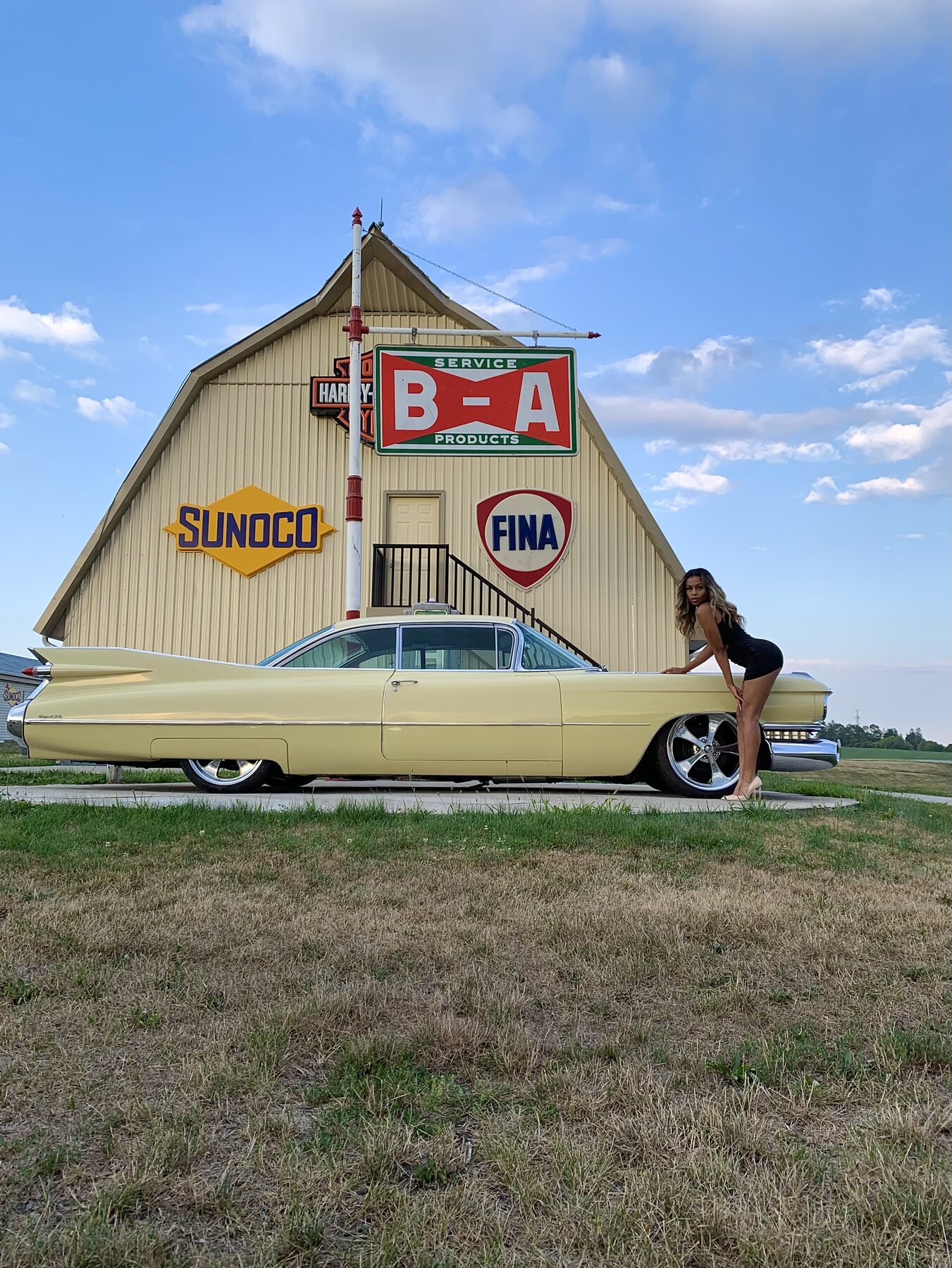 Tash Williamson in a full-length outdoor shot, bent forward at the waist with one hand braced on the hood of a vintage pastel-yellow finned Cadillac, wearing a fitted black sleeveless bodysuit and heels, parked in front of a retro service-station building covered in vintage Sunoco, Fina, and Harley-Davidson signage.