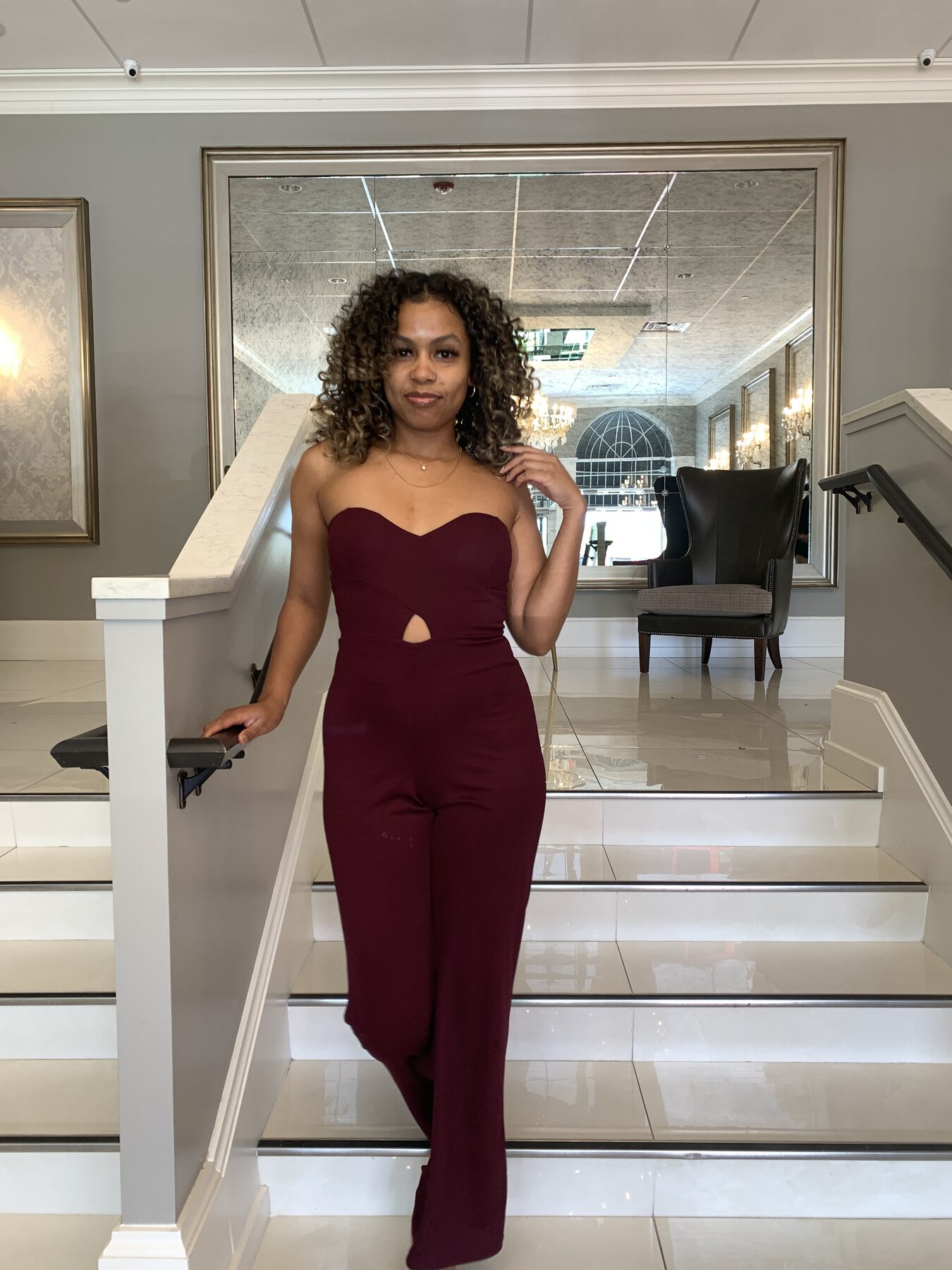 Tash Williamson in a full-length shot paused on a marble staircase in an upscale interior, leaning back against the white banister with one hand raised to her jaw and her elbow lifted, gazing off to the side with lips slightly parted, wide-leg burgundy jumpsuit with a sweetheart neckline and a keyhole cutout at the waist, curly hair with blonde balayage falling loose, other hand braced on the railing behind her.