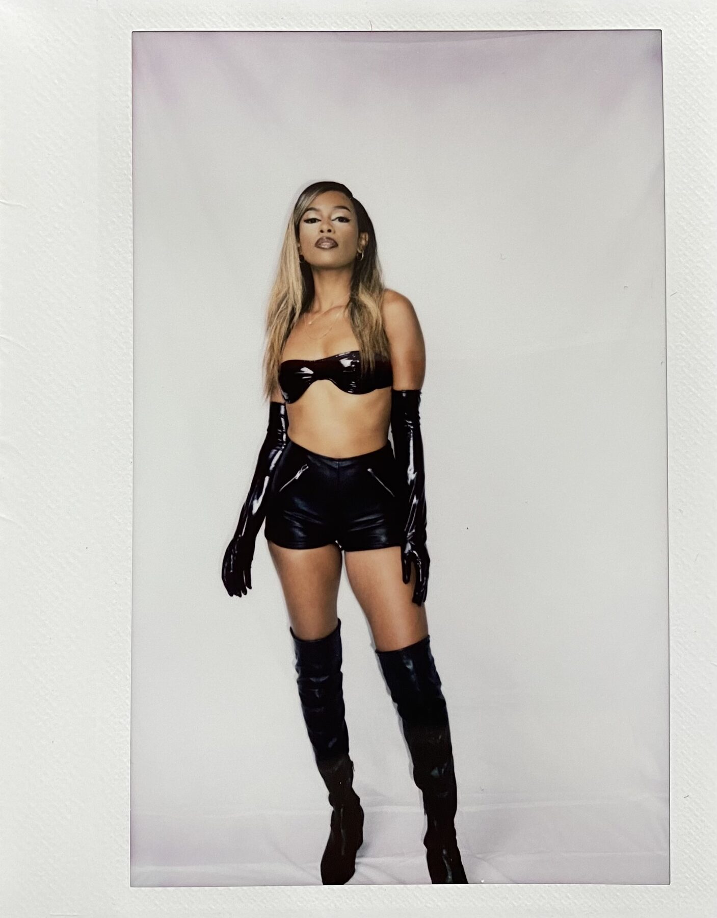 Tash Williamson in a full-length polaroid-framed studio shot, standing straight-on with arms relaxed at her sides, wearing the same black patent bandeau top, high-waisted pleather shorts, and long patent gloves as a companion frame, here shown head-to-toe with thigh-high patent boots and hair worn sleek and straight down her back.