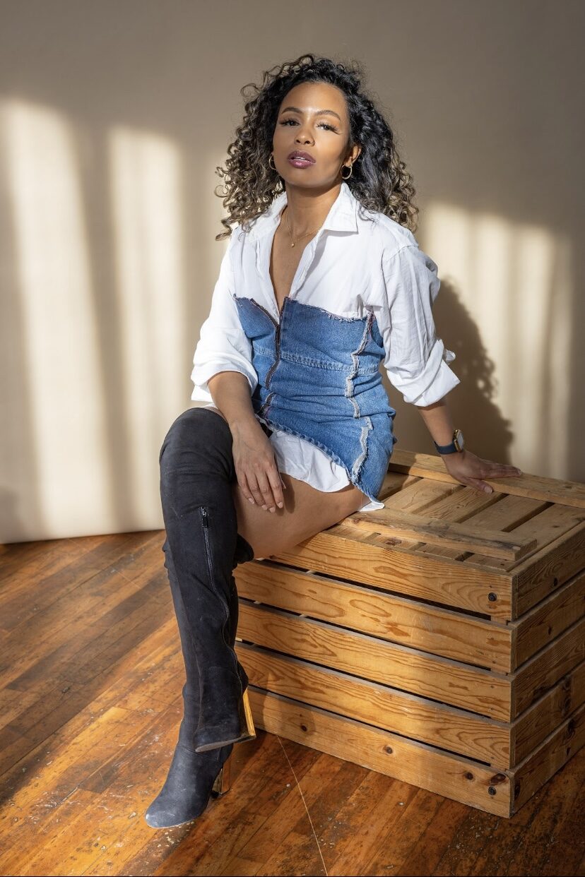 Tash Williamson seated upright on the wooden crate with legs crossed and black thigh-high boots on display, meeting the camera with a composed, closed-mouth gaze and the hint of a smirk, one hand resting on her lap and the other propped on the crate behind her, white shirt open over the raw-edge denim corset, curly balayage hair loose.