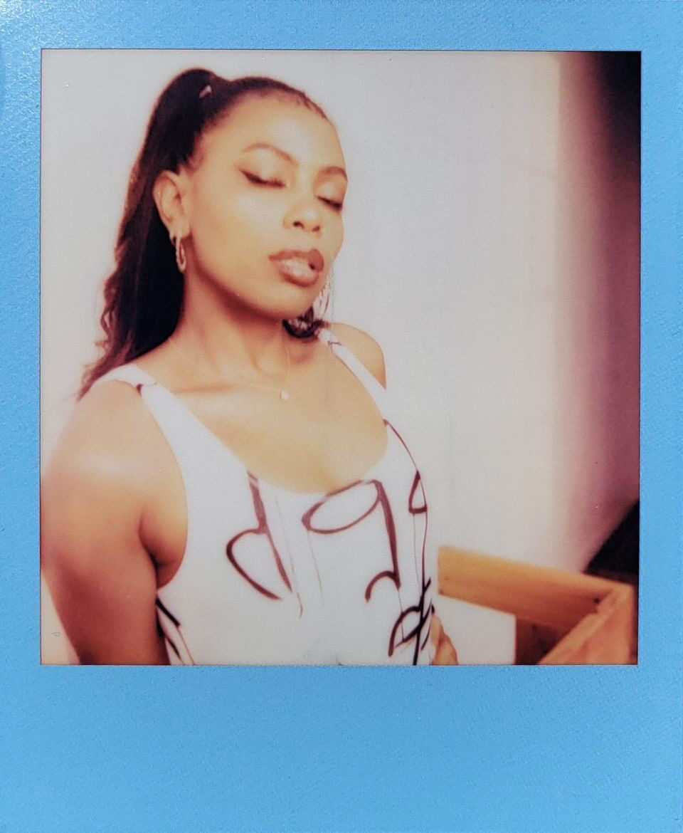 Tash Williamson in a Polaroid-style photograph with a blue border, a close medium shot with eyes closed and her chin tilted downward, wearing a white top or bodysuit with a dark abstract graphic print, hair pulled back into a sleek ponytail, softly lit indoors near a window.