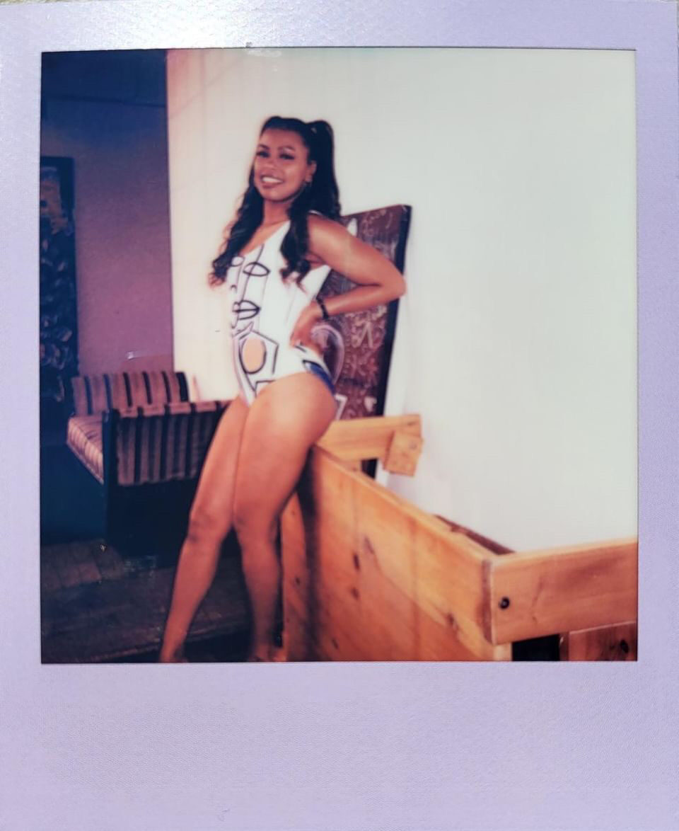 Tash Williamson in a Polaroid-style photograph with a purple border, a full-length front-facing shot leaning against a wooden crate with one hand on her hip, laughing with a wide open smile, wearing a white swimsuit with a bold graphic print, ponytail with loose curls framing her face, a striped bench and dark artwork visible in the background.