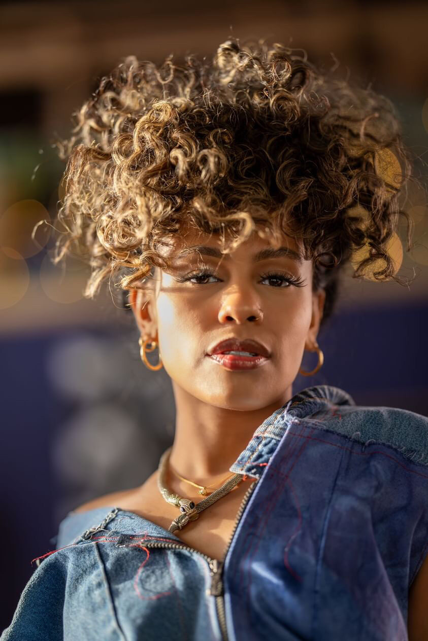 Tash Williamson standing upright and facing the camera directly in a home kitchen, hands resting together near the tied waistband of her teal crochet pants, midriff bared through the cutout halter top revealing a belly-button piercing, loose wavy curls framing her face, a window with blinds and a dish rack visible behind her.